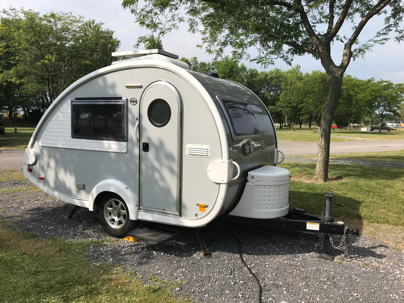 A sleek, grey travel trailer RV rental sits on a gravel campsite, surrounded by green trees. Ready for adventure.