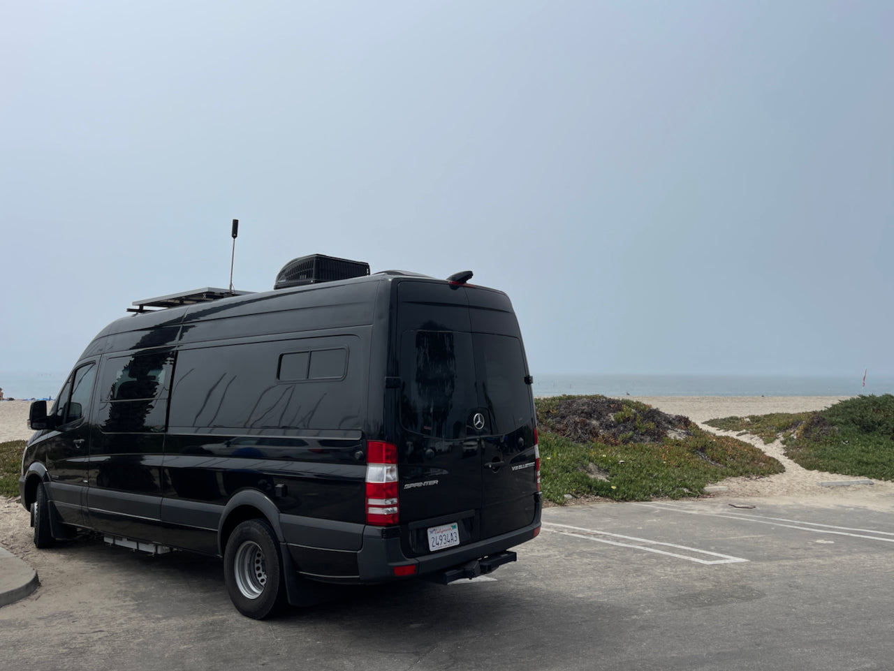 Black Sprinter rv rental parked at a serene beachside lot. Ocean, sandy path, and hazy sky visible, perfect for coastal adventures.