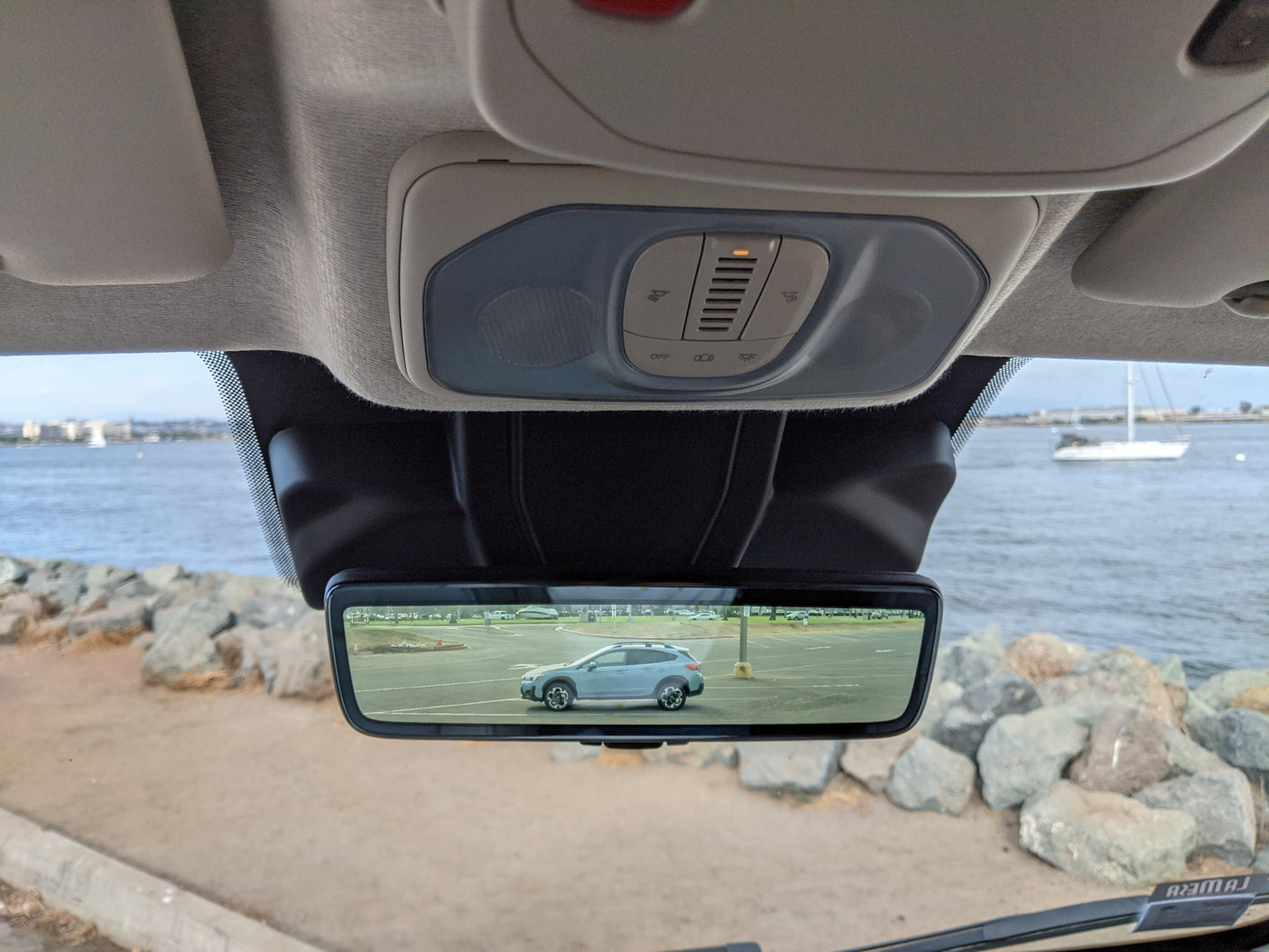 View from an RV rental, showing the interior overhead console and a digital mirror reflecting a light blue car parked by the waterfront.