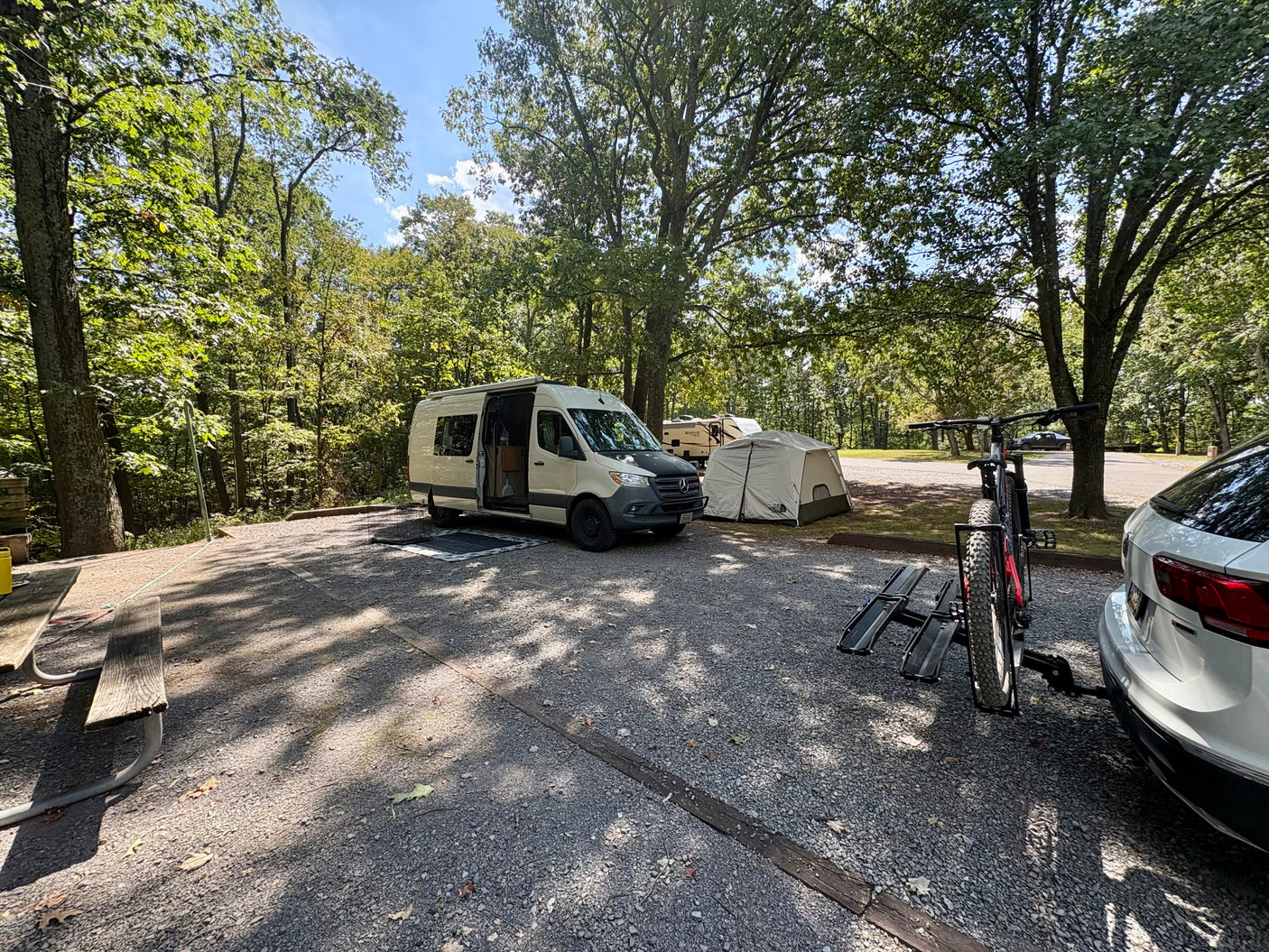 An RV rental camper van, tent, and mountain bike are set up at a shaded, wooded campsite for a relaxing trip.