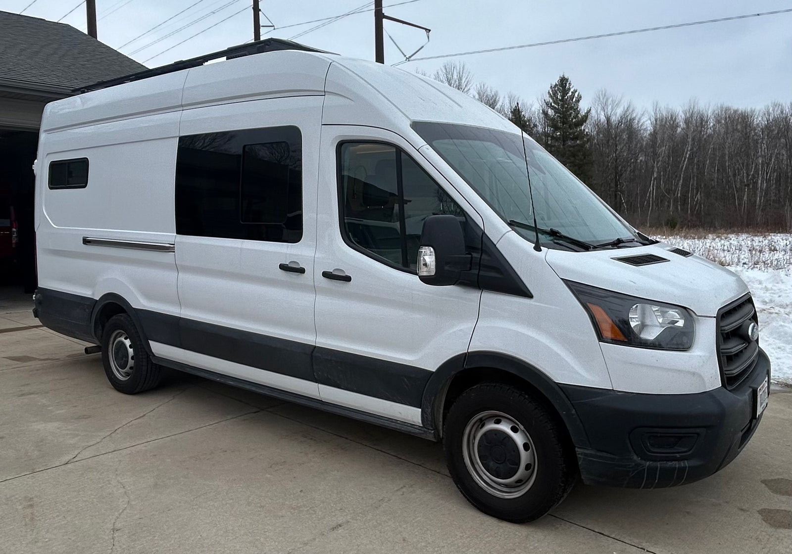 White Ford Transit camper van, perfect for an RV rental adventure. Parked on a concrete driveway with a snowy, wooded background.