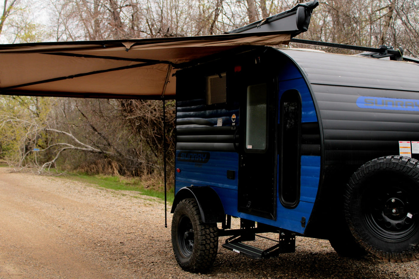 A blue and black teardrop camper RV rental with an extended awning, parked on a gravel path amidst trees. Ready for adventure.