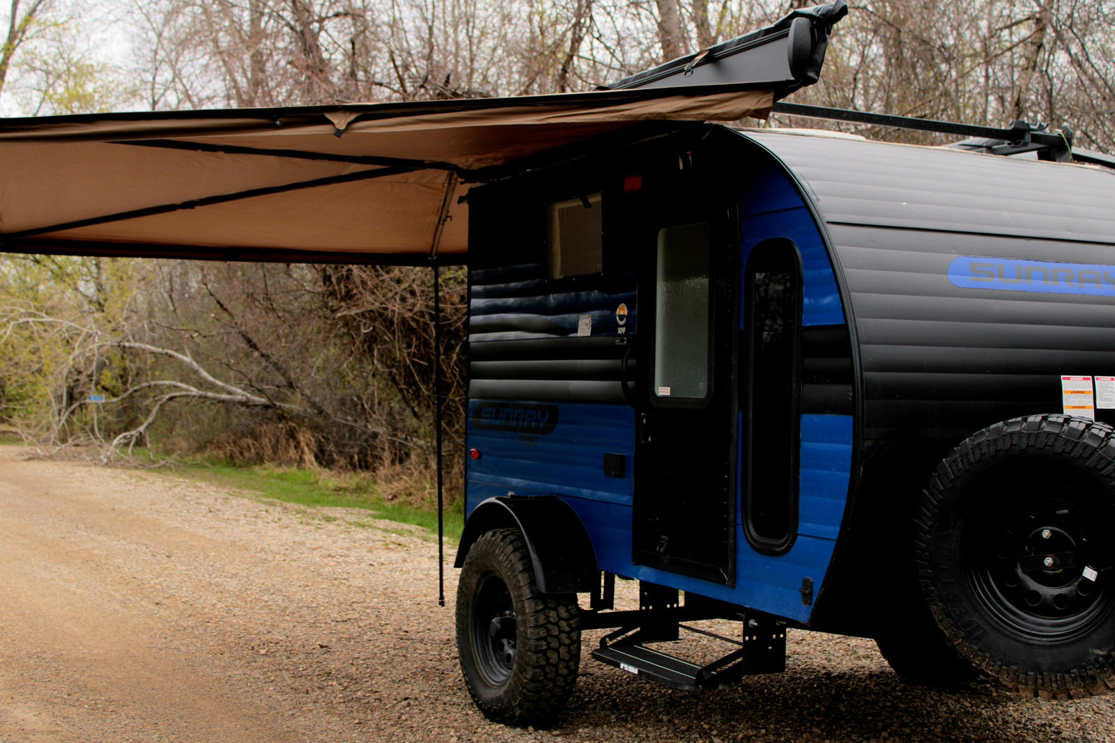 A blue and black teardrop camper RV rental with an extended awning, parked on a gravel path amidst trees. Ready for adventure.