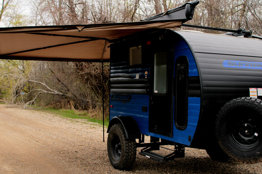 A blue and black teardrop camper RV rental with an extended awning, parked on a gravel path amidst trees. Ready for adventure.