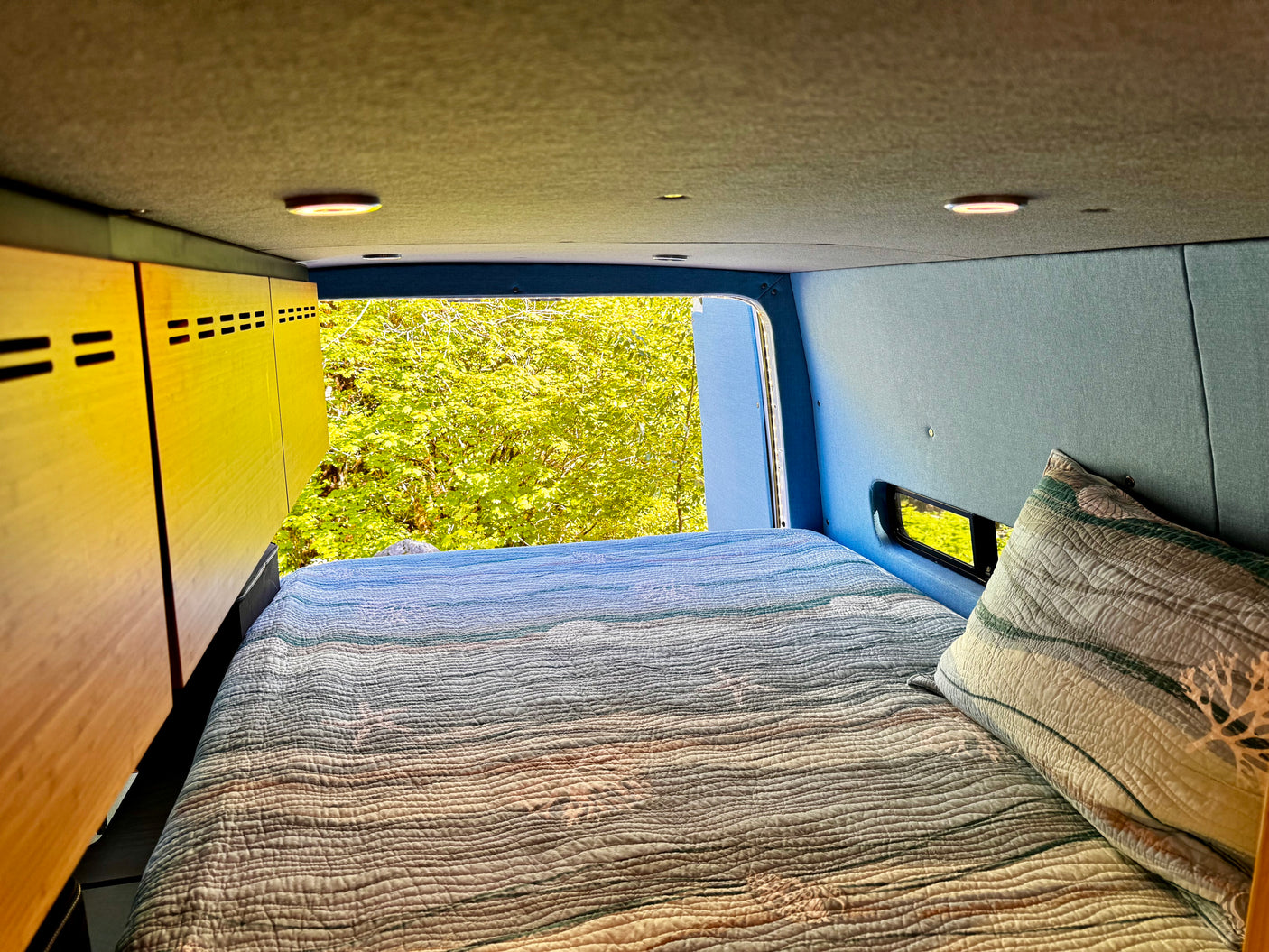 Inside this cozy RV rental, a comfy quilted bed faces lush green nature views through the open back.