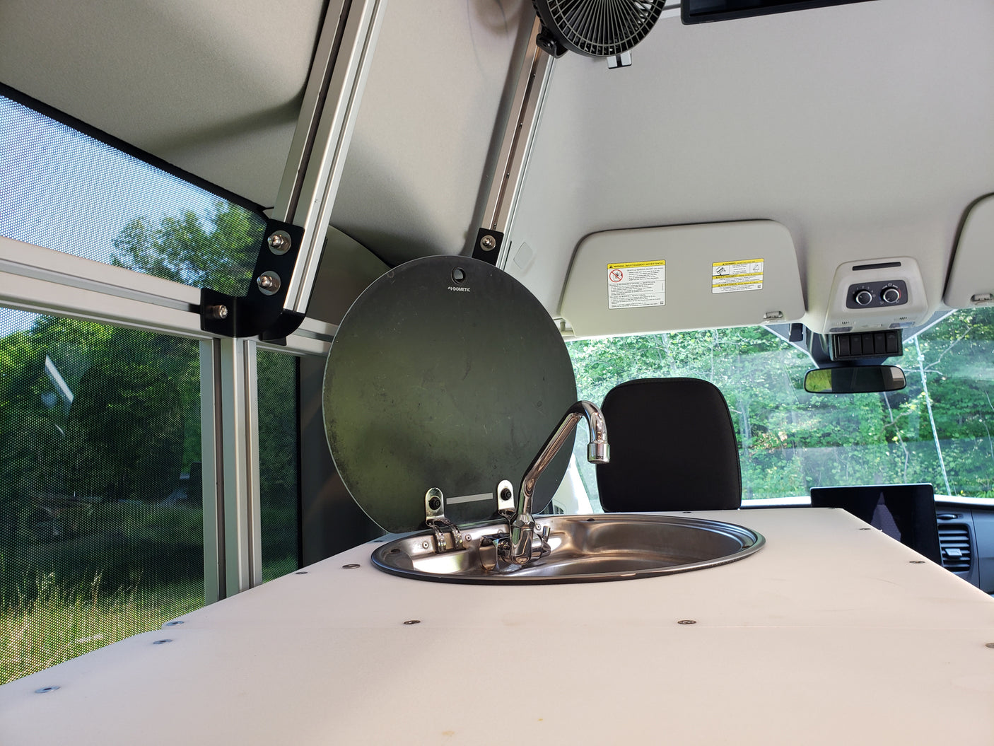 This RV rental interior showcases a compact kitchen area with a stainless steel sink, faucet, and a light countertop. A mesh window overlooks green trees outside.