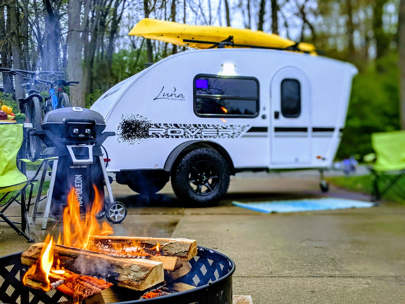 An RV rental camping scene features a Luna Rover teardrop trailer with kayak, bikes, grill, and a warm campfire.