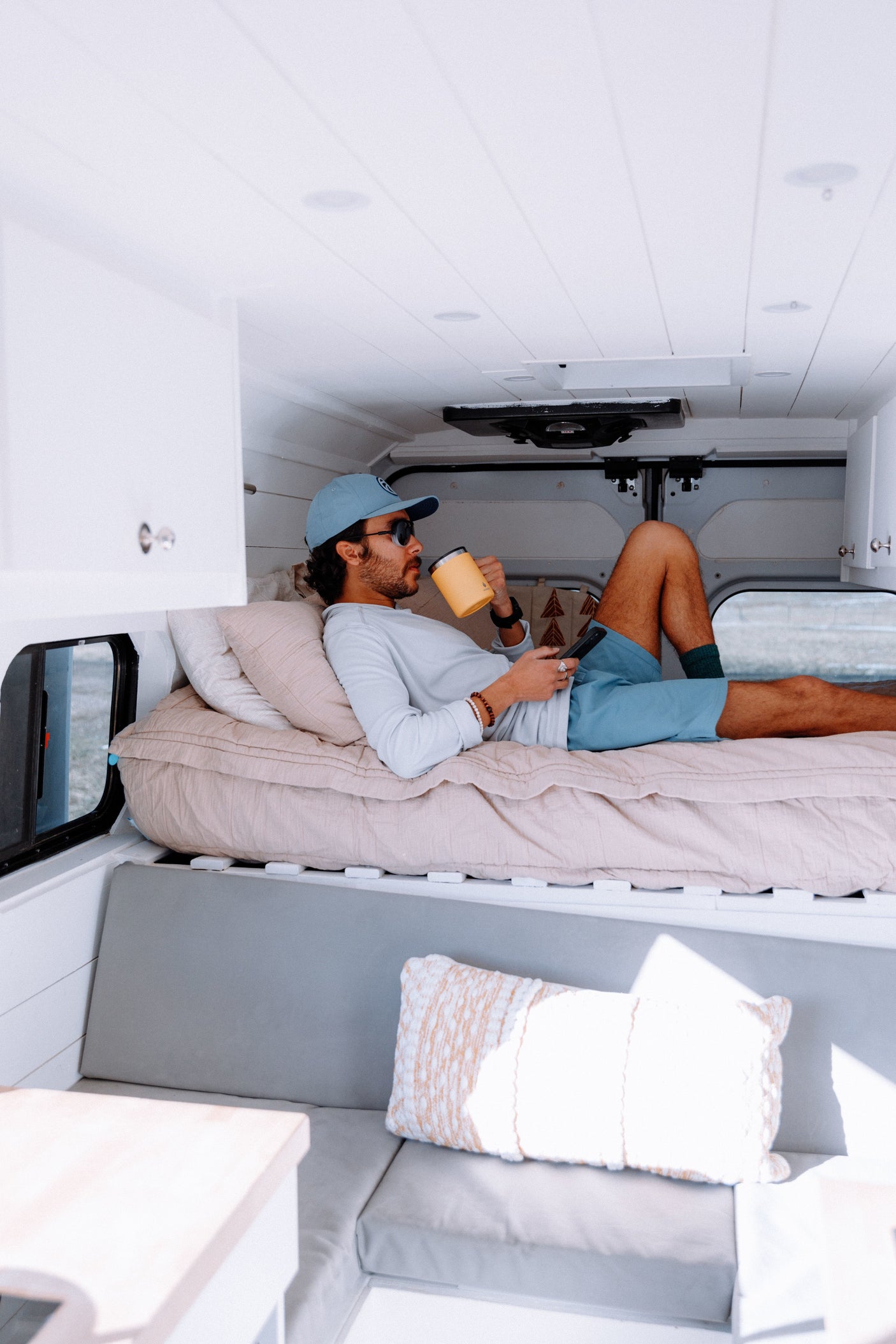 A man relaxes on a bed inside a clean, modern RV rental, holding a yellow mug & phone. A cozy interior for van life.