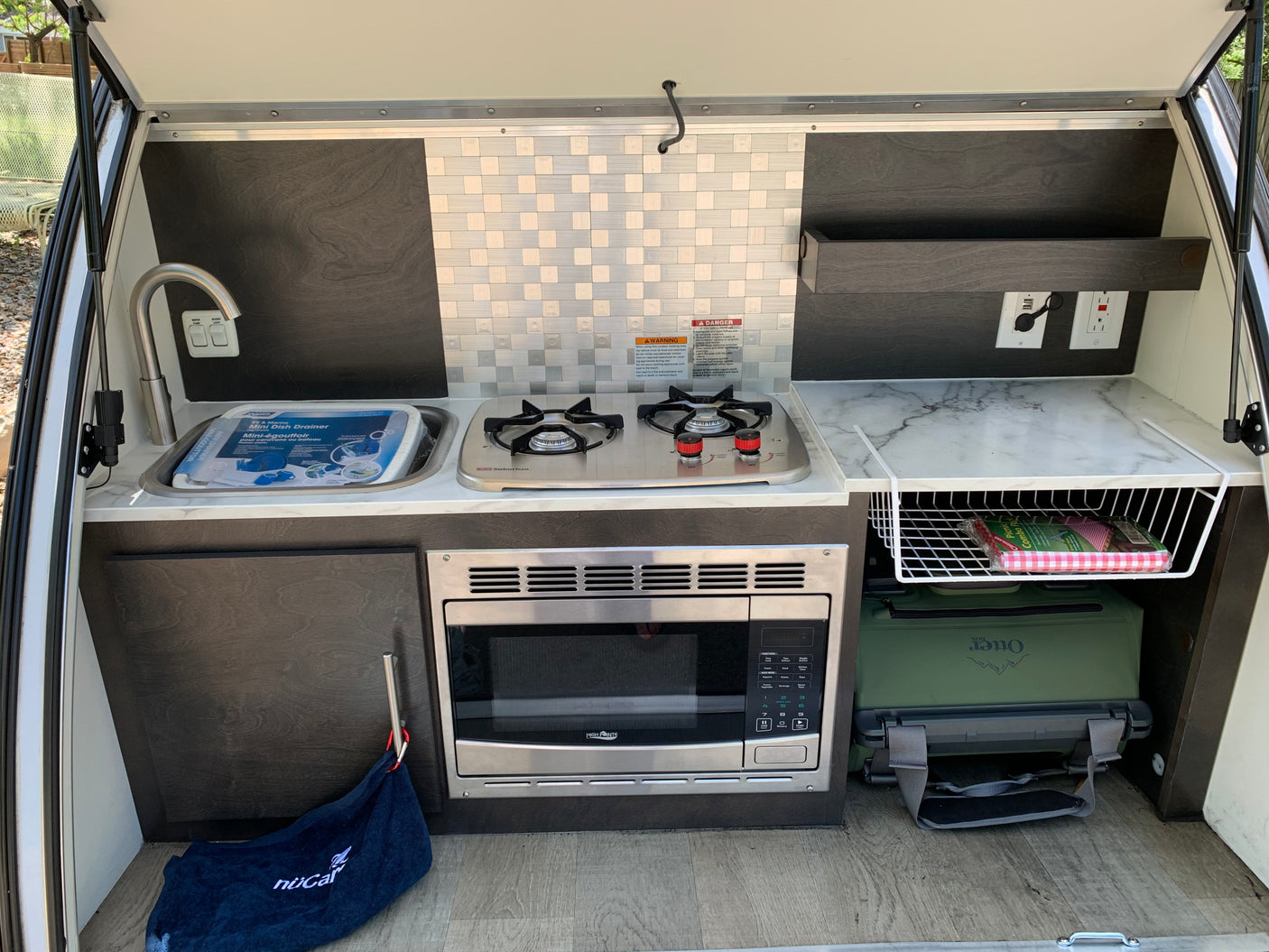 Outdoor galley kitchen in an RV rental or teardrop trailer, featuring a sink, two-burner stove, microwave, and cooler for glamping convenience.