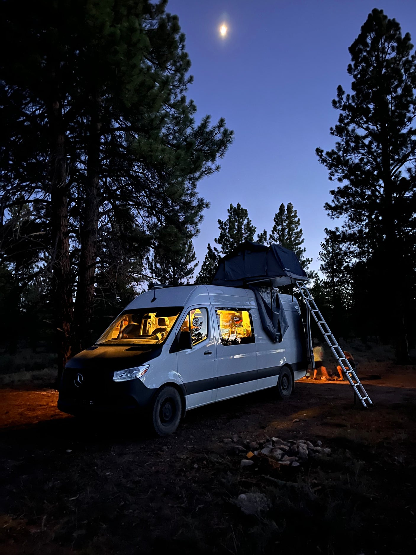 Night camping with an RV rental: a lit camper van, rooftop tent, and people under a moonlit sky.