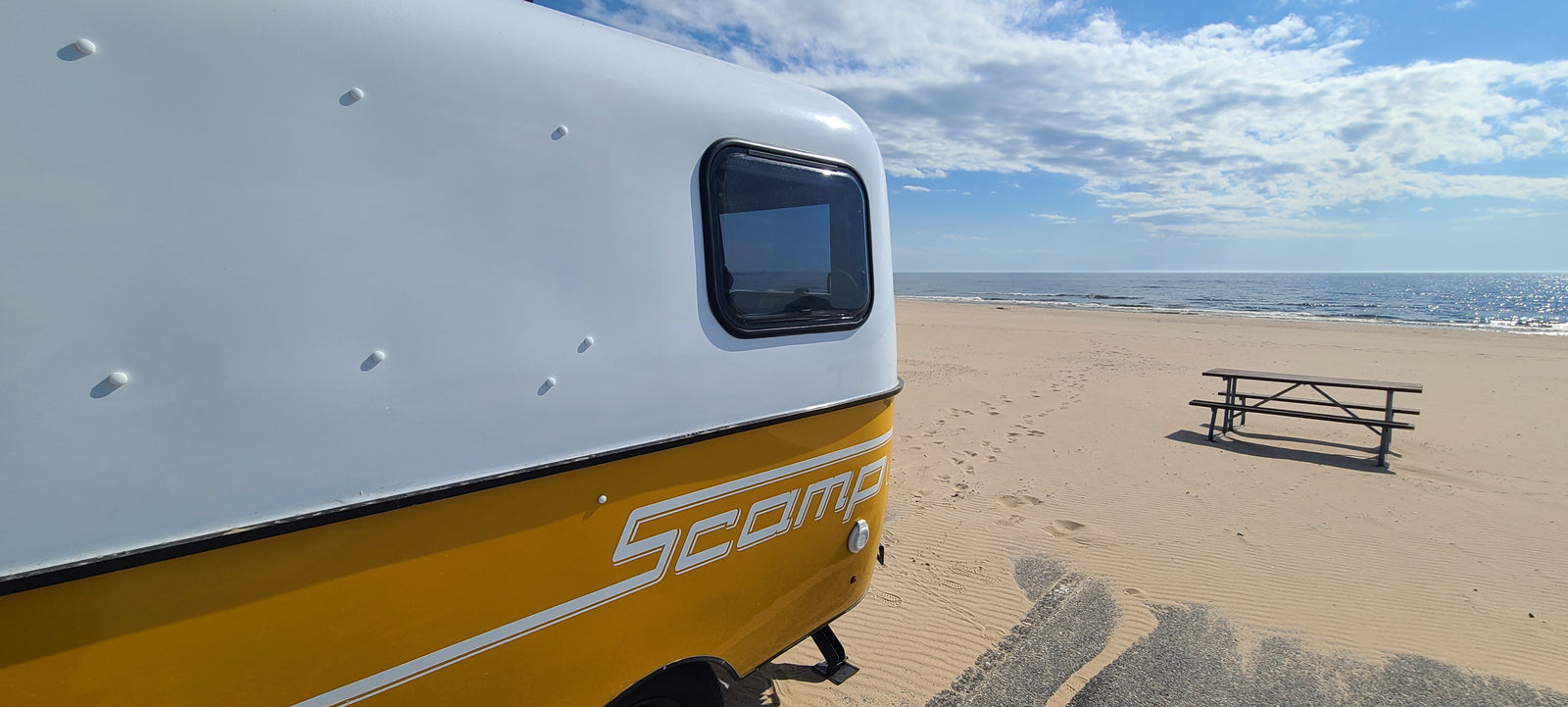 Scamp RV rental on a serene sandy beach, overlooking the sparkling ocean under a blue sky, with a picnic bench nearby.