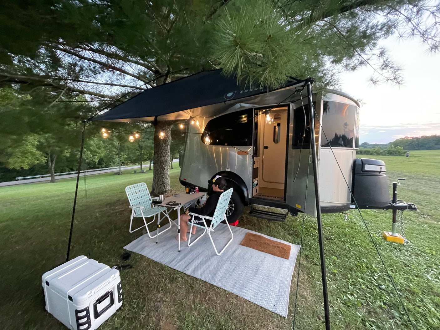 Enjoying an RV rental. A person dines outside a modern travel trailer under an awning with string lights at dusk.