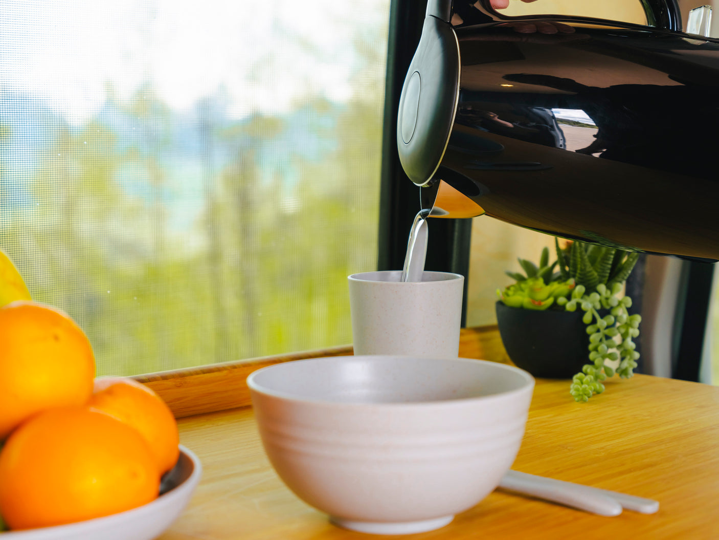 Pouring water from a black kettle into a cup inside an RV rental. Fresh oranges and a plant sit on the wooden counter by a window.