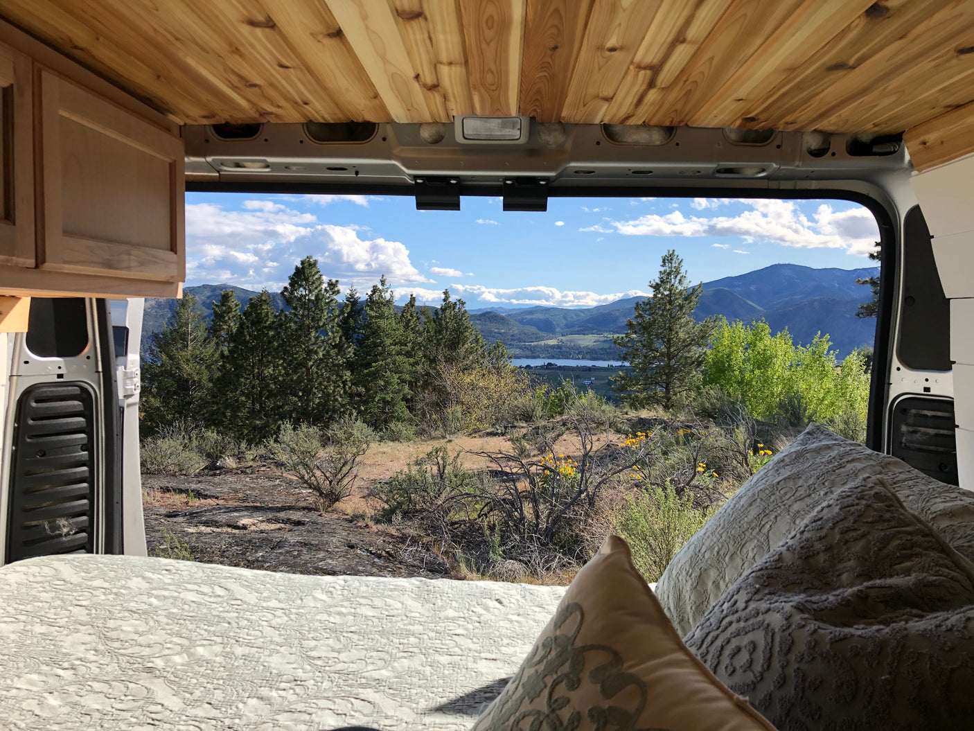 RV rental: Inside a campervan, a bed offers a scenic view of mountains, a lake, and trees under blue skies.
