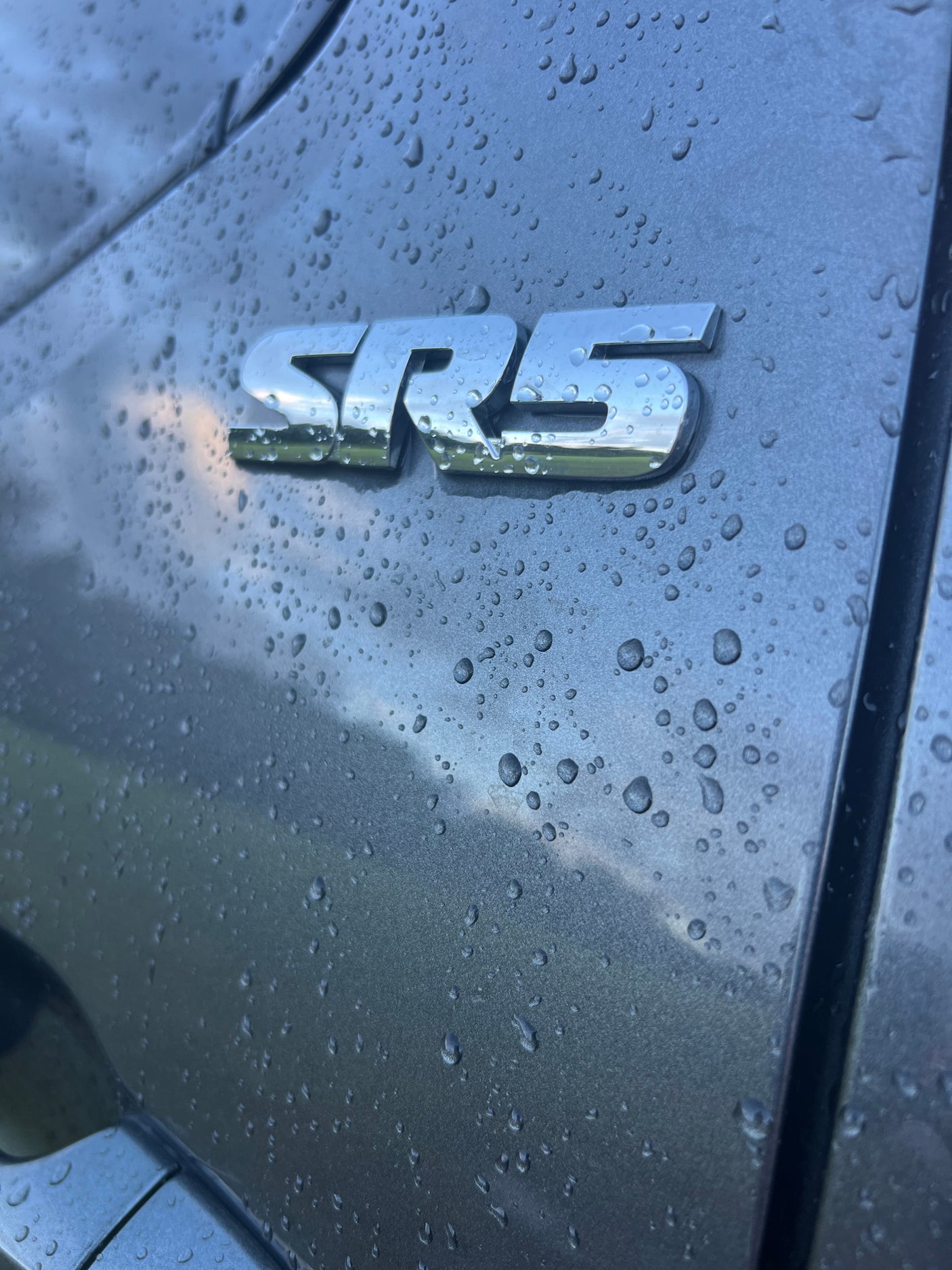Close-up: Rain-covered grey vehicle with a shiny 'SRS' emblem reflecting cloudy skies. Ideal for an RV rental ad.