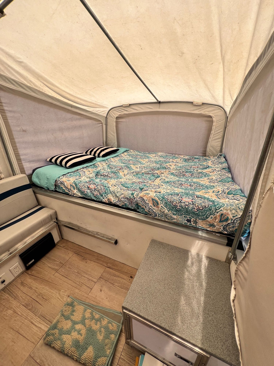 is set up in a serene forest campsite.|Pop-up camper RV rental set up in a wooded campsite with a child walking near a fire pit
