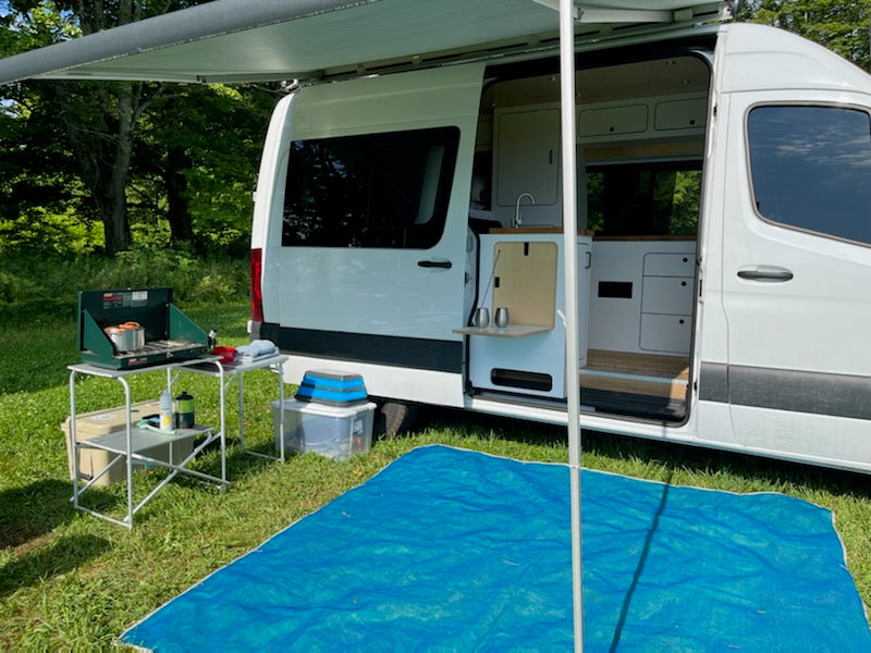 RV rental camper van with open door revealing a modern kitchen, awning extended for outdoor cooking setup.