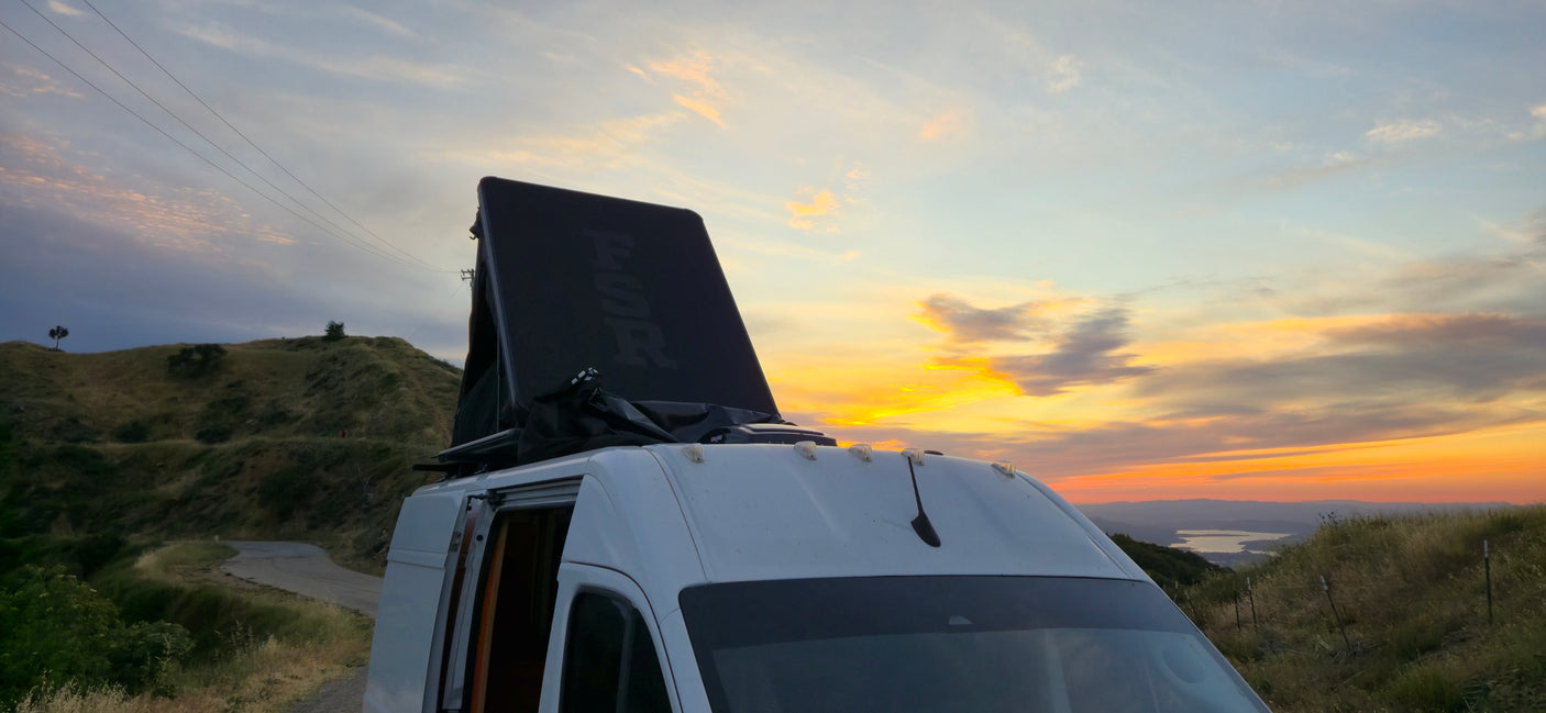 An RV rental with a deployed rooftop tent captures a vibrant sunset over hills and a distant lake. Scenic road trip!