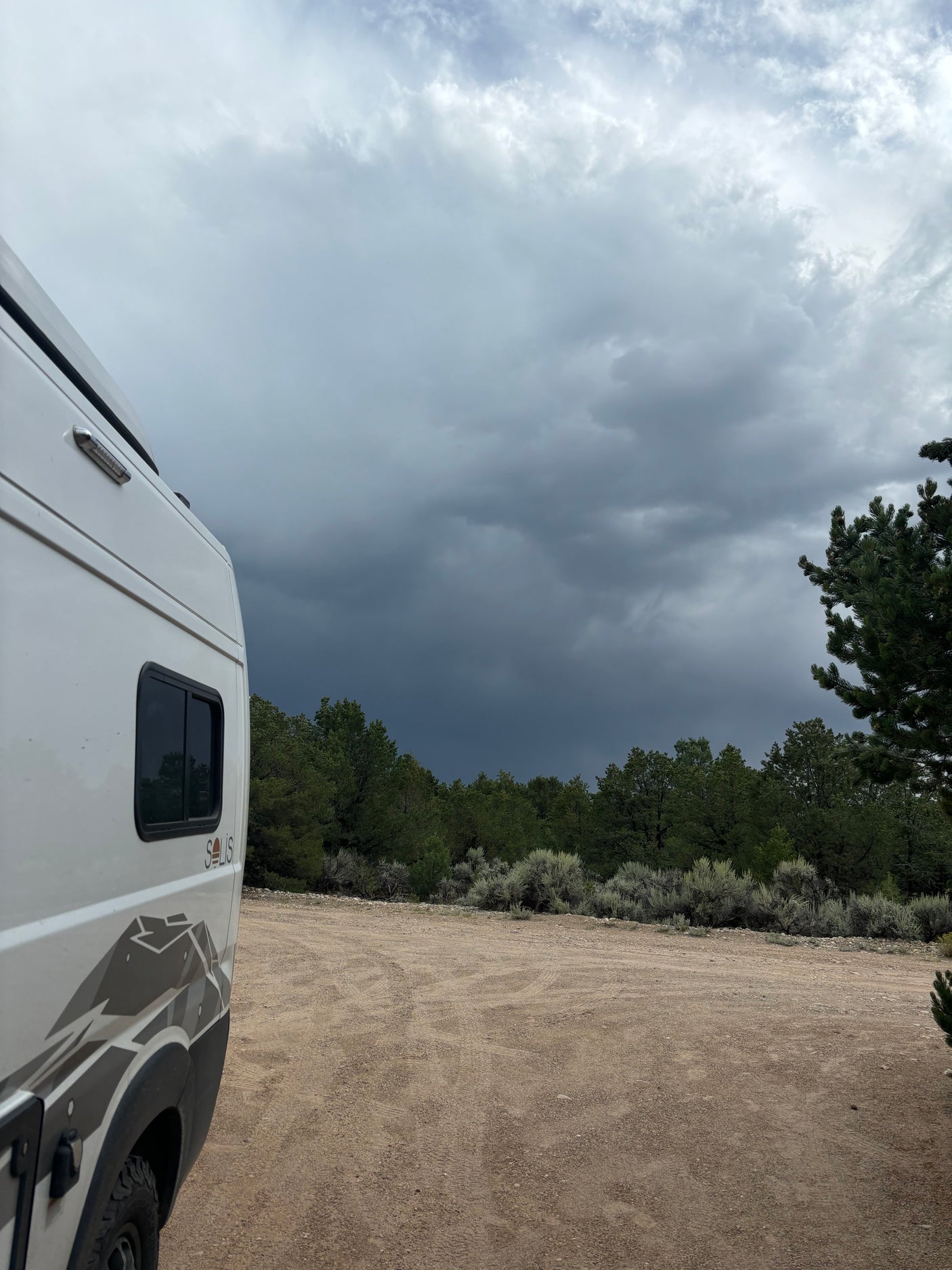 RV rental (Winnebago Solis) on a dirt road amidst trees, under a dramatic, stormy sky. Perfect for remote outdoor adventures.