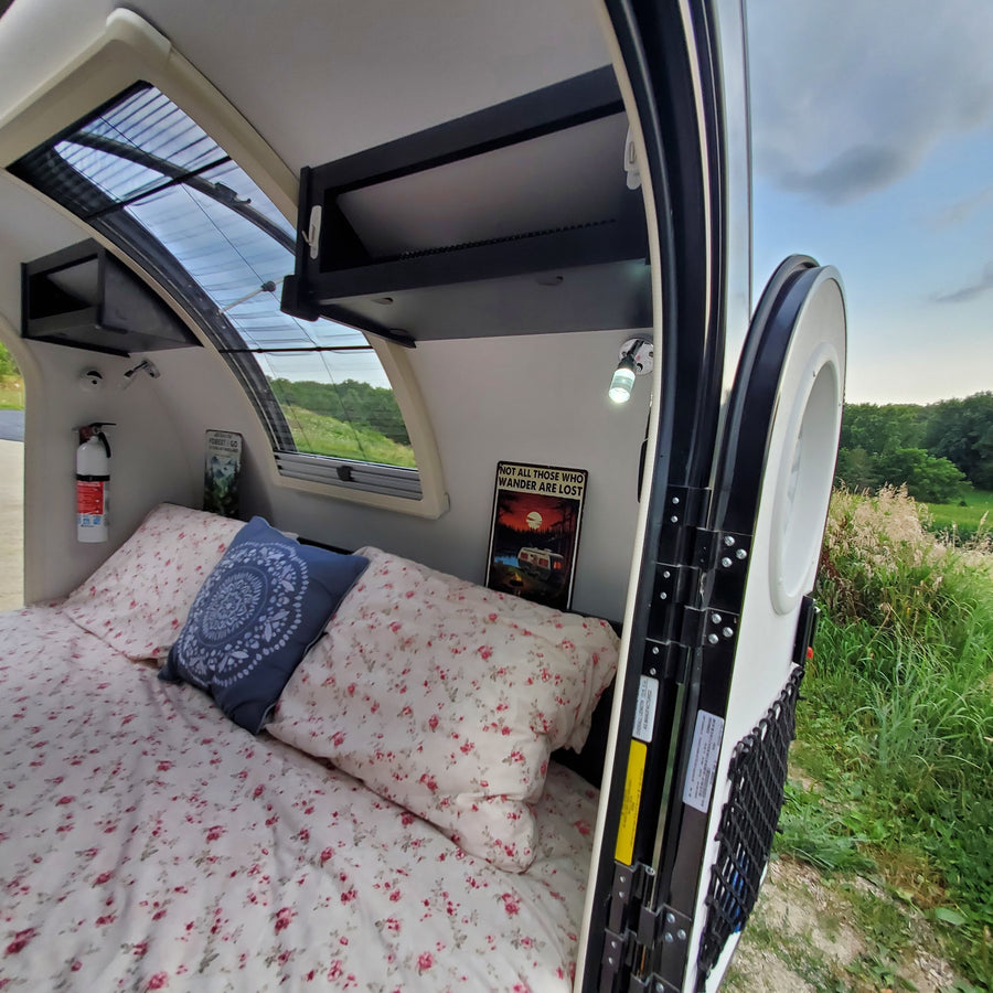 Step inside this cozy RV rental camper, featuring a comfortable bed, overhead storage, and an open door to nature.