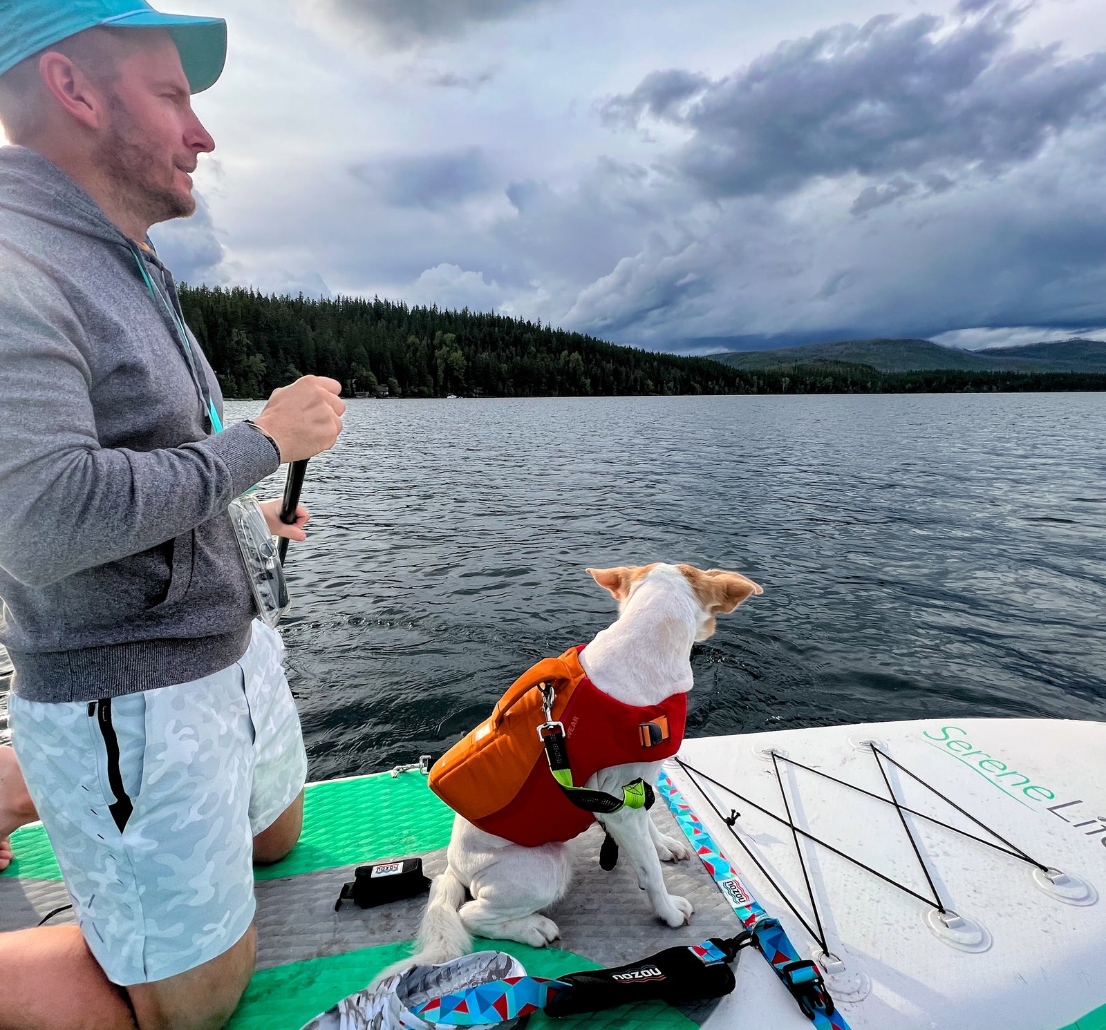 A man and his dog on a paddleboard, enjoying a lake day, perfect after an RV rental journey. Mountains and clouds in background.