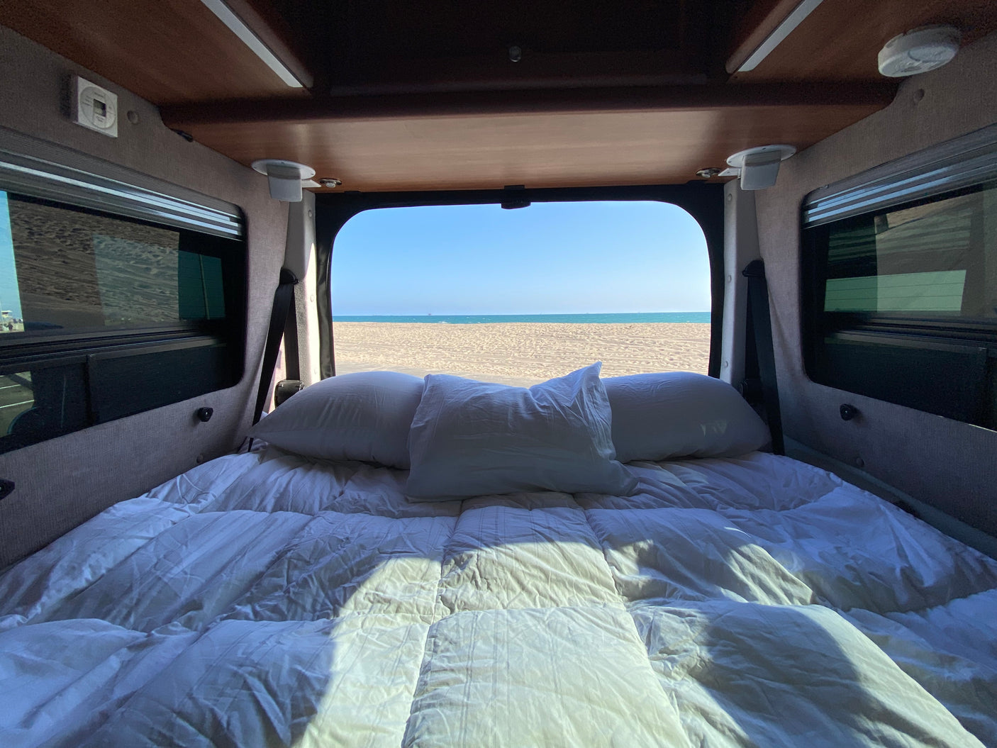Cozy rv rental bed with white linens and pillows, framed by the open back, reveals a stunning sandy beach & clear ocean view.