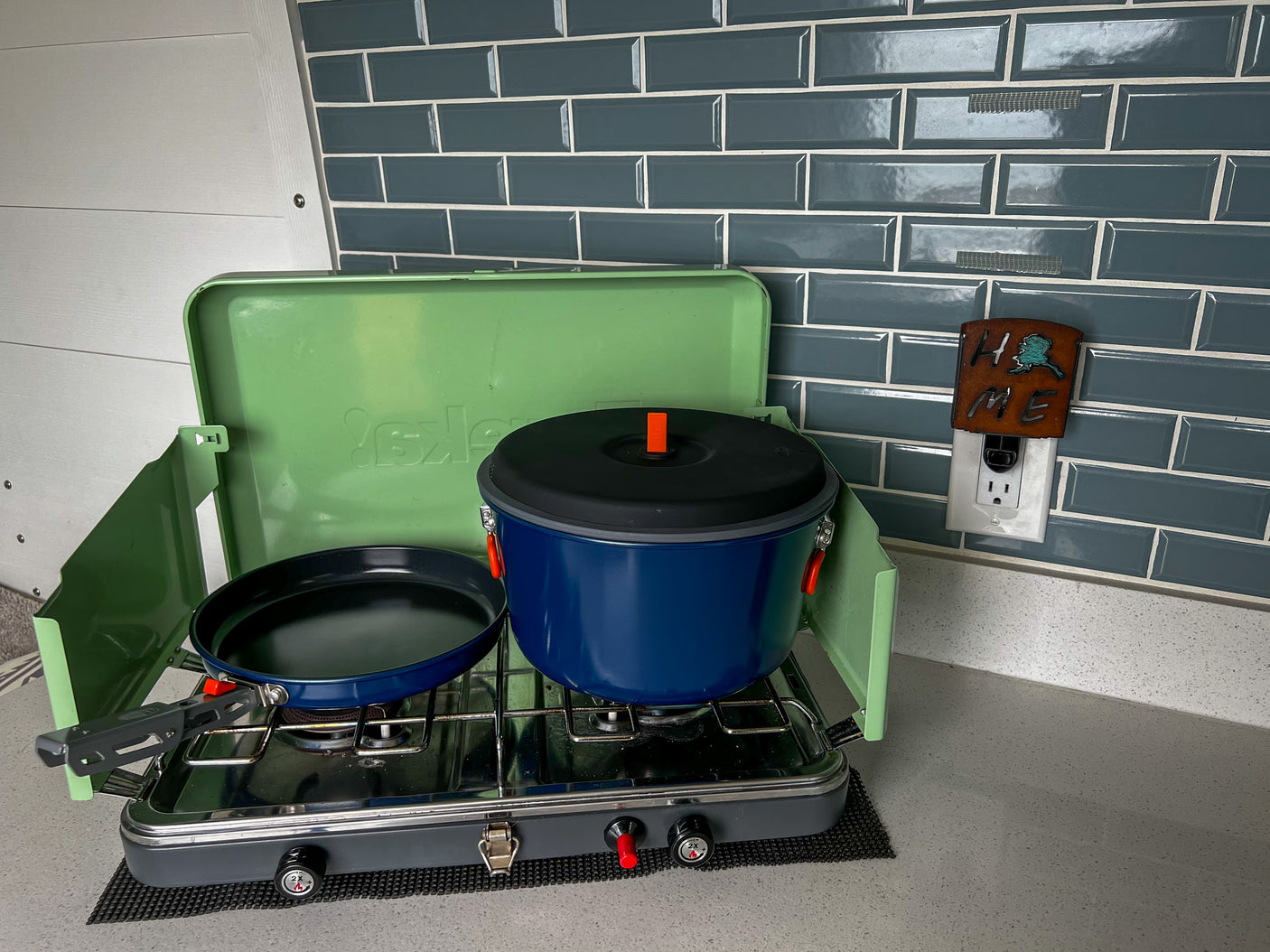 Compact RV rental kitchen: green portable camping stove with a blue pot & black pan on a gray counter.