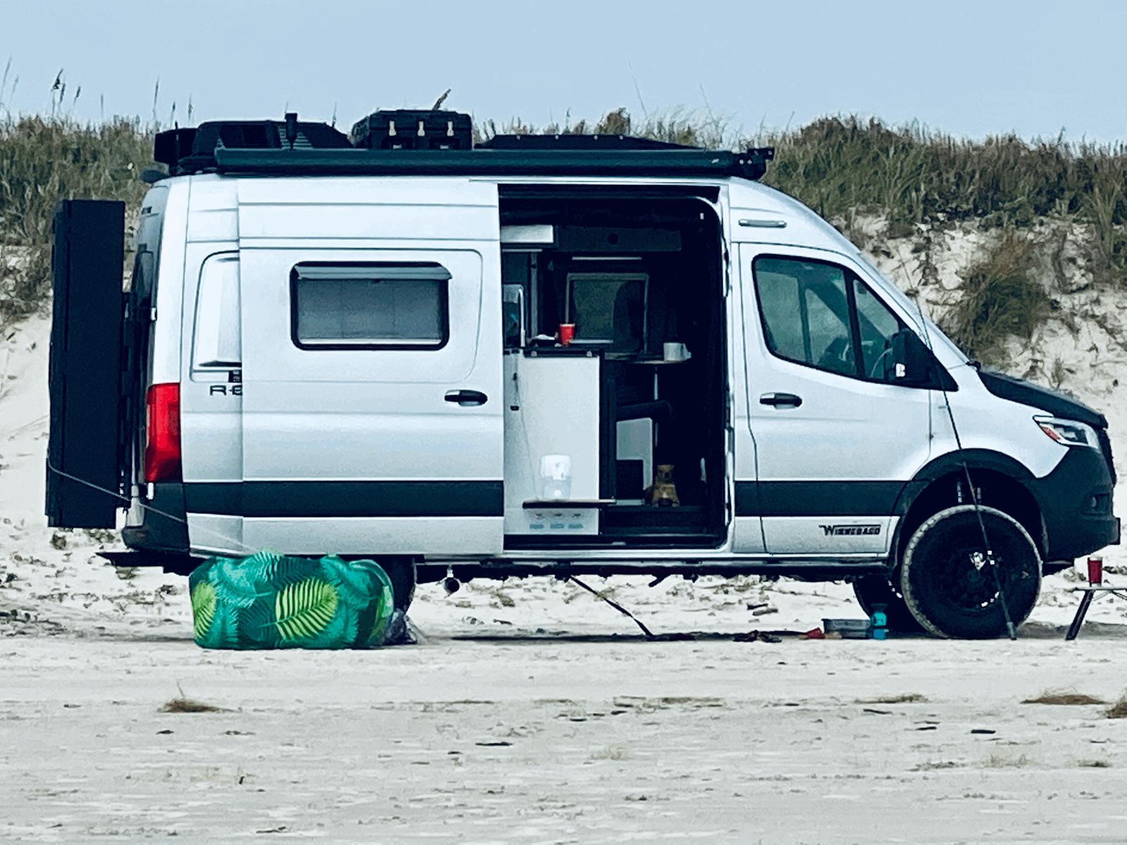 Winnebago RV rental camper van parked on a sandy beach. Its side door is open, showing a cozy interior ready for adventure.