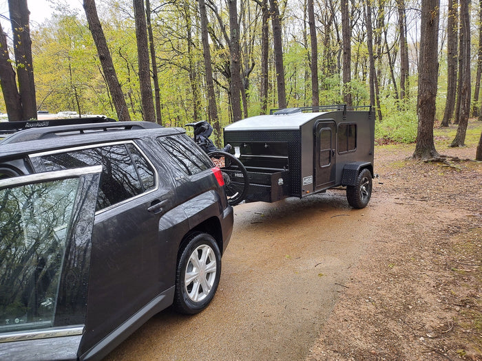 RV rental: dark SUV towing a Hiker Trailer camper & bike. Ready for adventure in a wet, lush forest setting.
