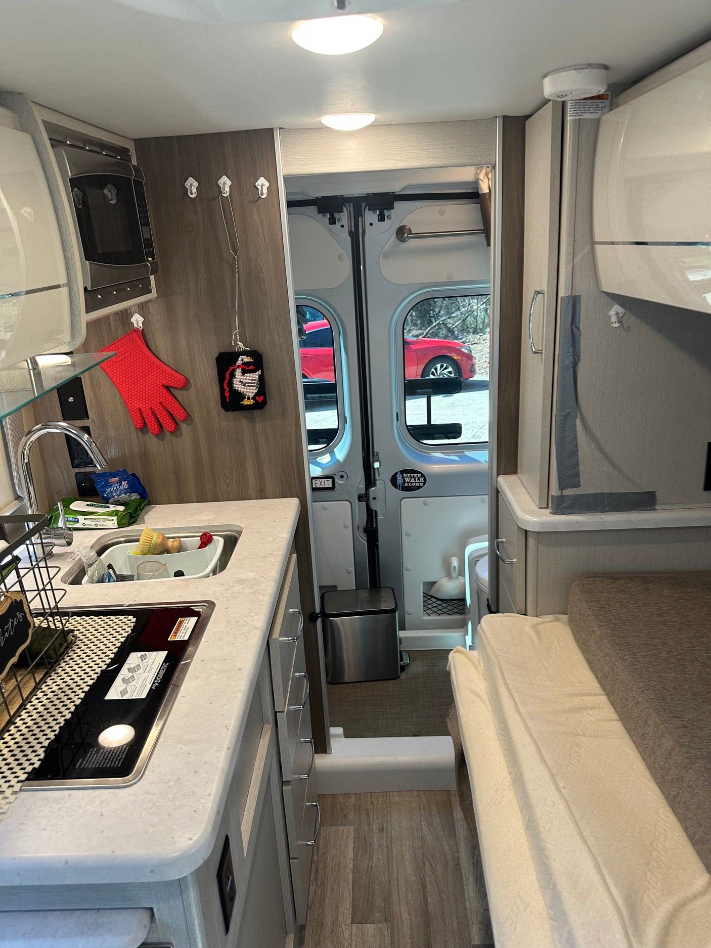 Inside a cozy RV rental: kitchenette with sink, stove, and microwave, plus a bed and open rear doors to a car outside.