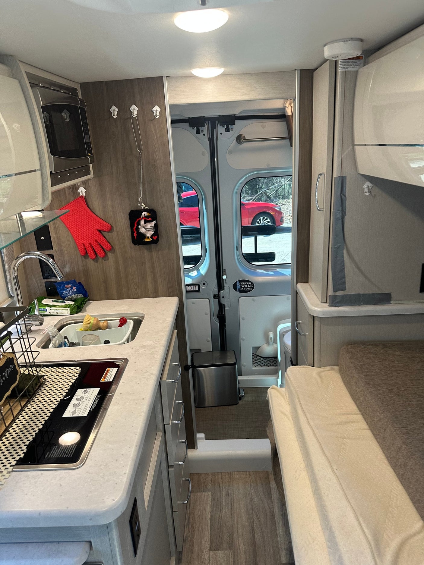 Inside a cozy RV rental: kitchenette with sink, stove, and microwave, plus a bed and open rear doors to a car outside.