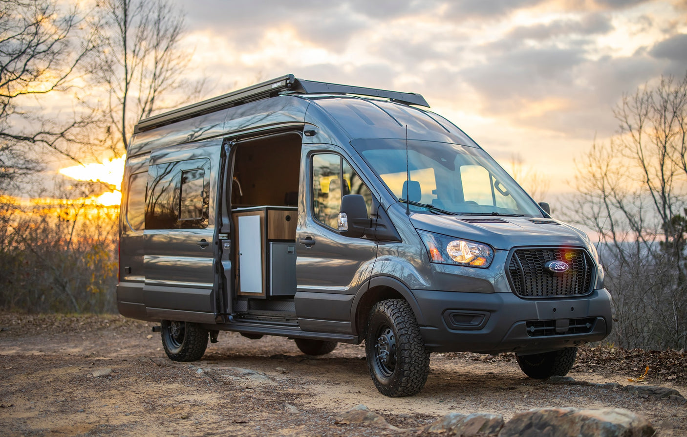 Grey Ford Transit rv rental camper van parked on a dirt path with an open door showing the modular interior.|Inside a modern RV rental van conversion: organized storage