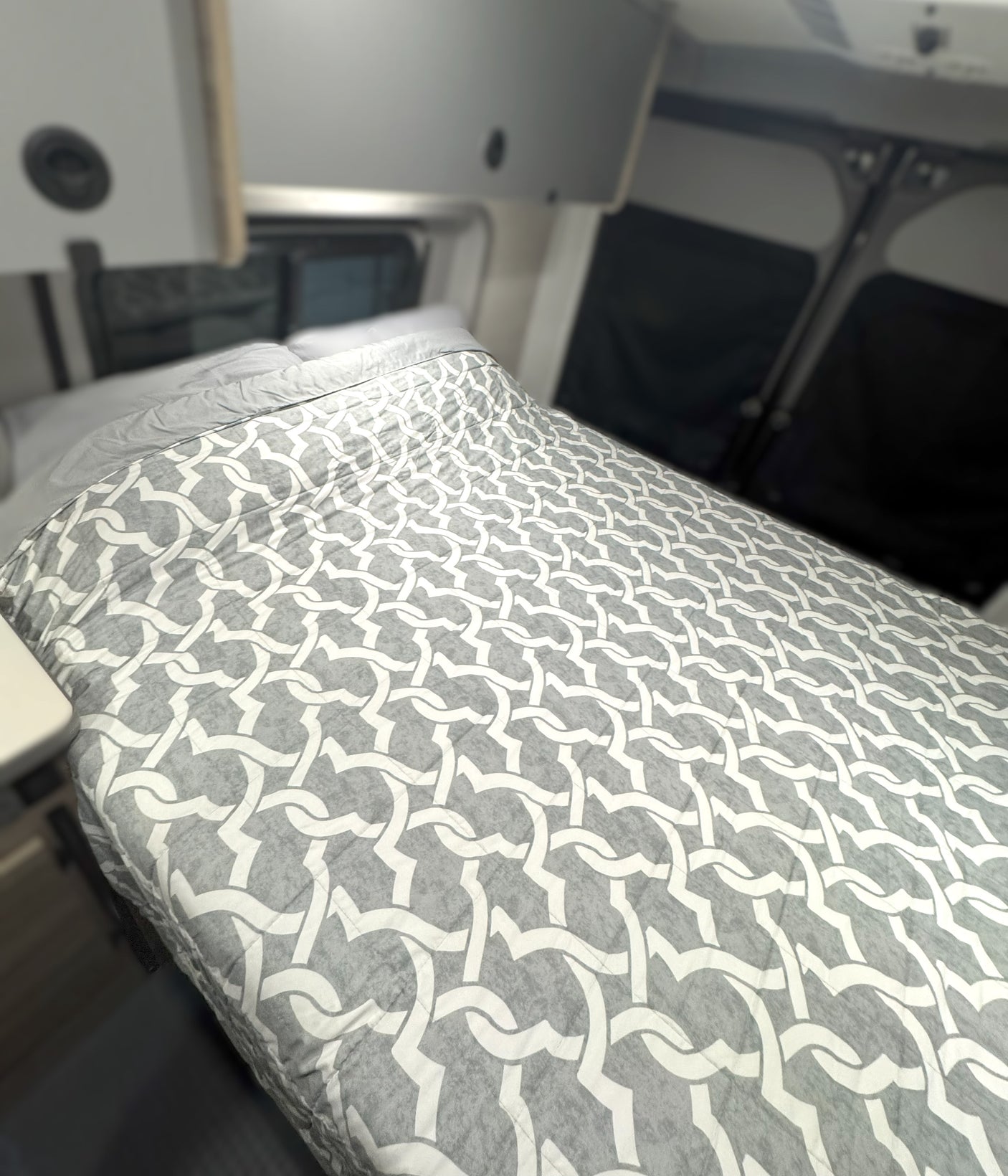 Cozy bed with a grey & white patterned comforter inside an RV rental. Overhead cabinets & window covers visible.