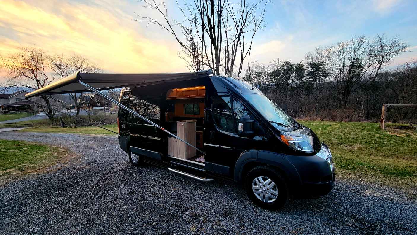 A black RV rental van with an extended awning, parked on a gravel drive at sunset. Interior visible, ready for adventures.