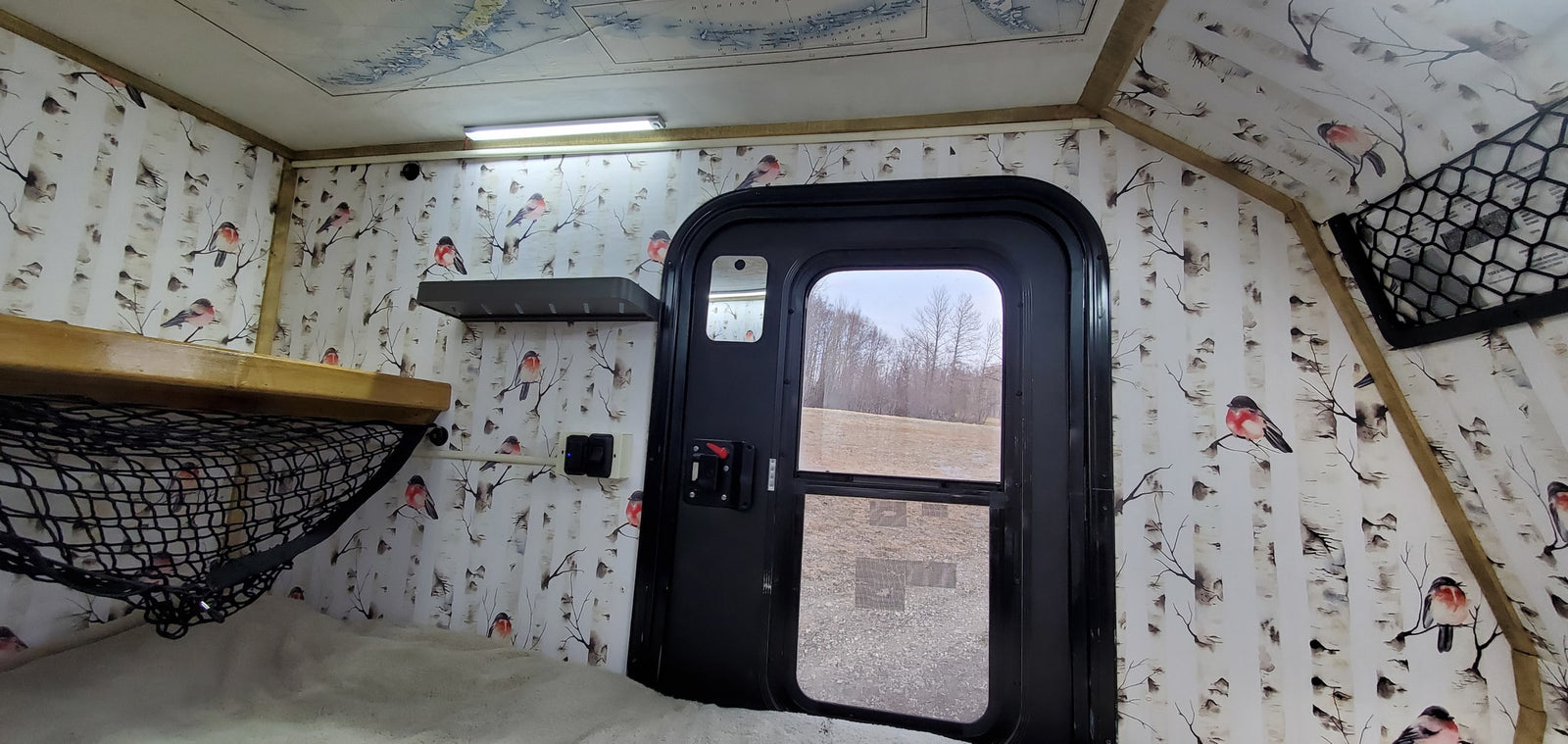 Cozy RV rental interior featuring a bed, storage net, and birch tree/bird decor. View out the door to fields.