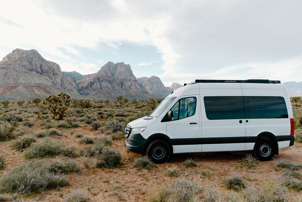 White Sprinter rv rental parked in a vast desert landscape, featuring rugged, layered mountains and desert brush under a cloudy sky.