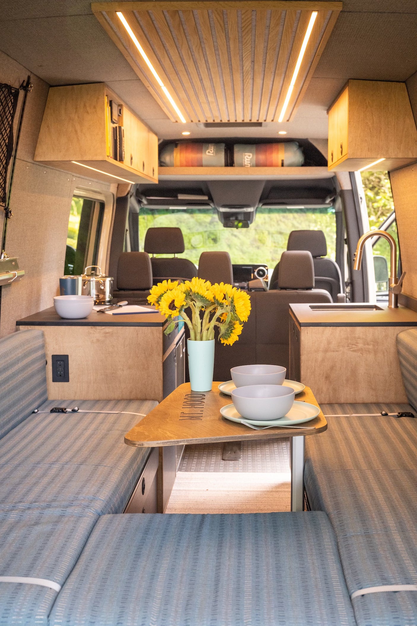 This RV rental interior features a stylish kitchen, dining area with sunflowers, and comfortable seating, ideal for mobile living.