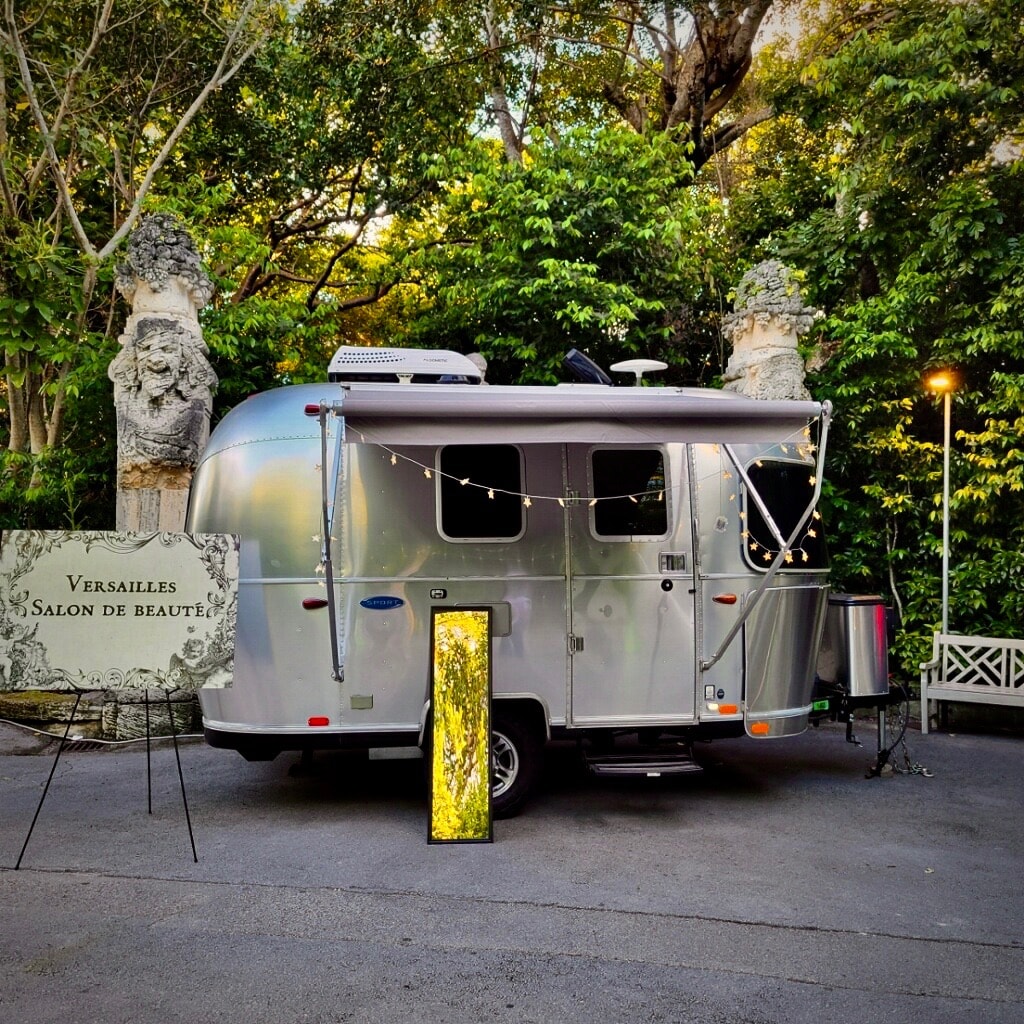 Airstream rv rental transformed into a 'Versailles Salon de Beauté' with awning, lights, & glowing display in a lush garden.