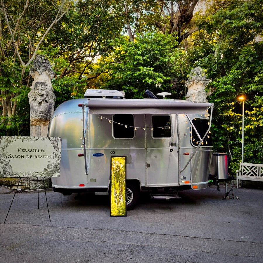 Airstream rv rental transformed into a 'Versailles Salon de Beauté' with awning, lights, & glowing display in a lush garden.
