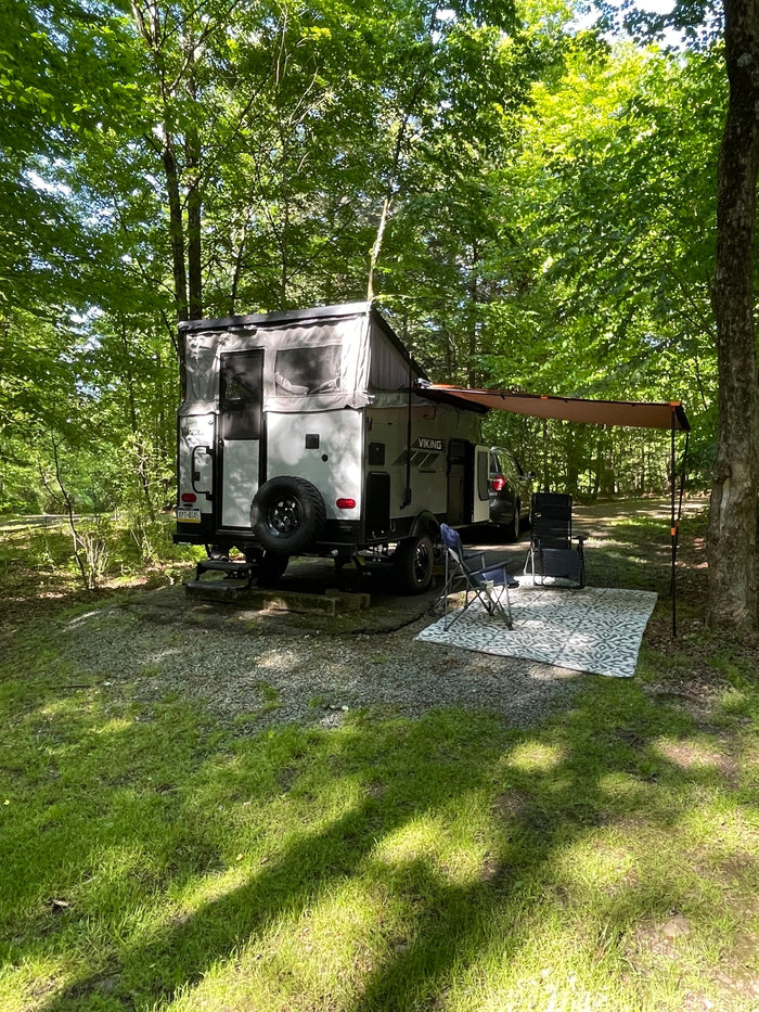 Enjoying a serene wooded campsite, this RV rental offers an extended awning, chairs, and rug for outdoor relaxation.