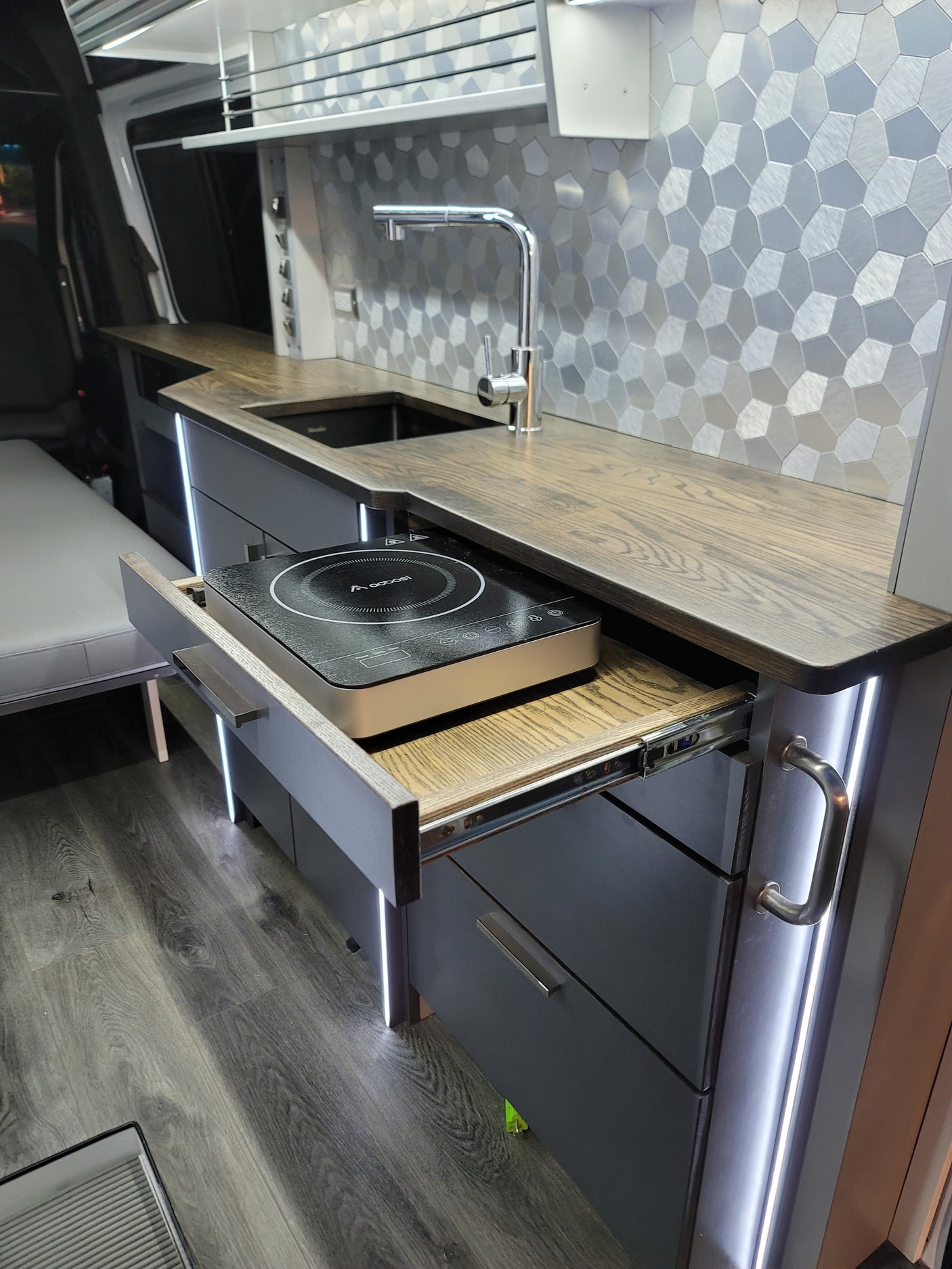 Sleek RV rental interior: modern kitchen with wood counters, sink, pull-out induction cooktop, and accent lighting.