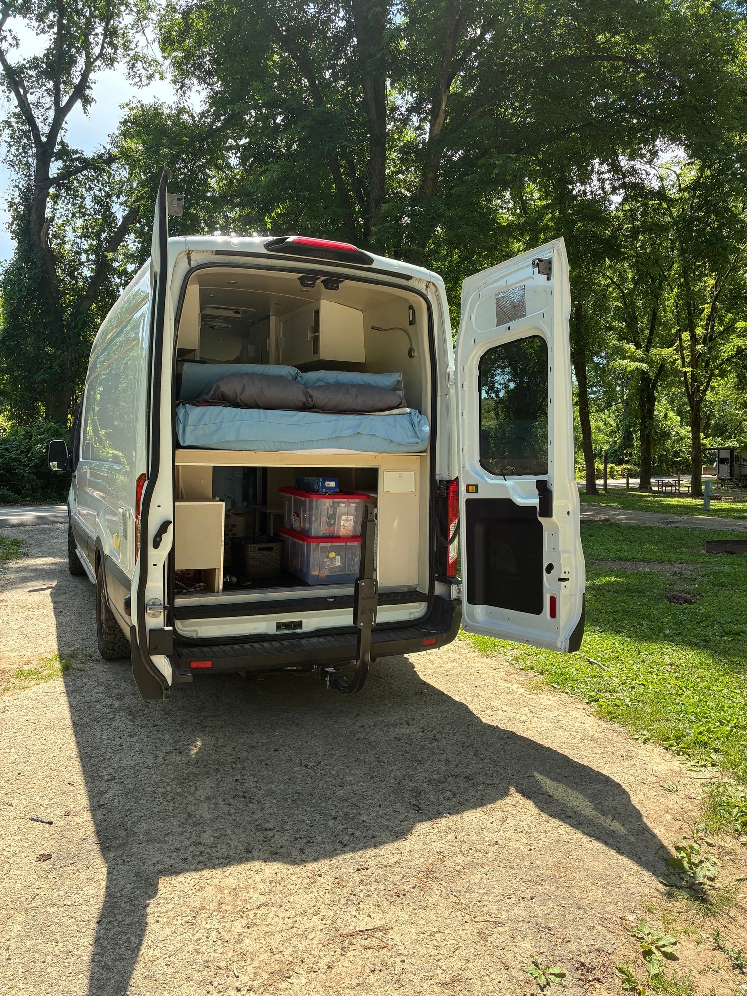 Back of a white RV rental camper van with open doors, showing interior bed and storage, at a sunny campsite.