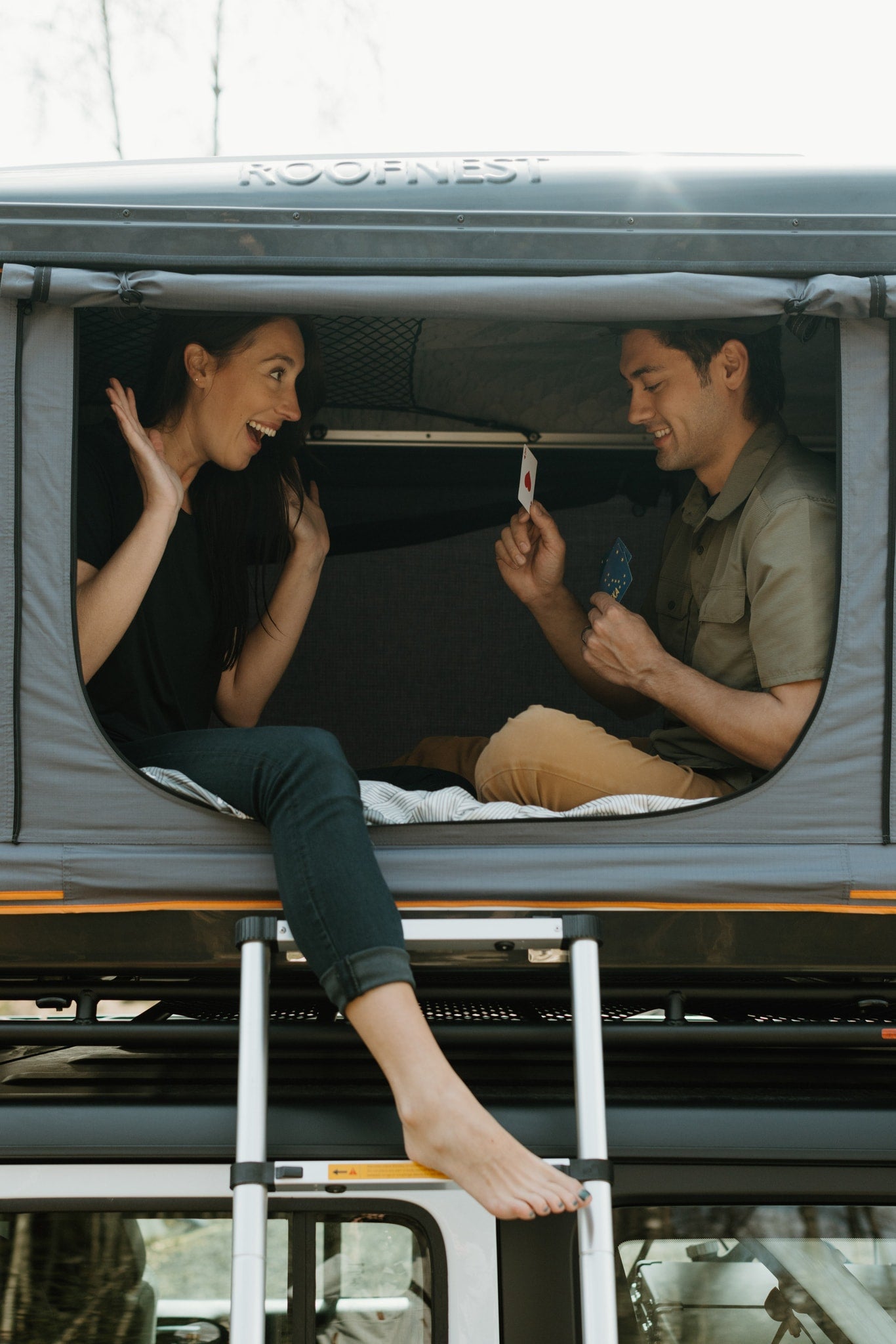 A happy couple enjoys cards in a gray Roofnest roof tent mounted on an RV rental, with a ladder visible.