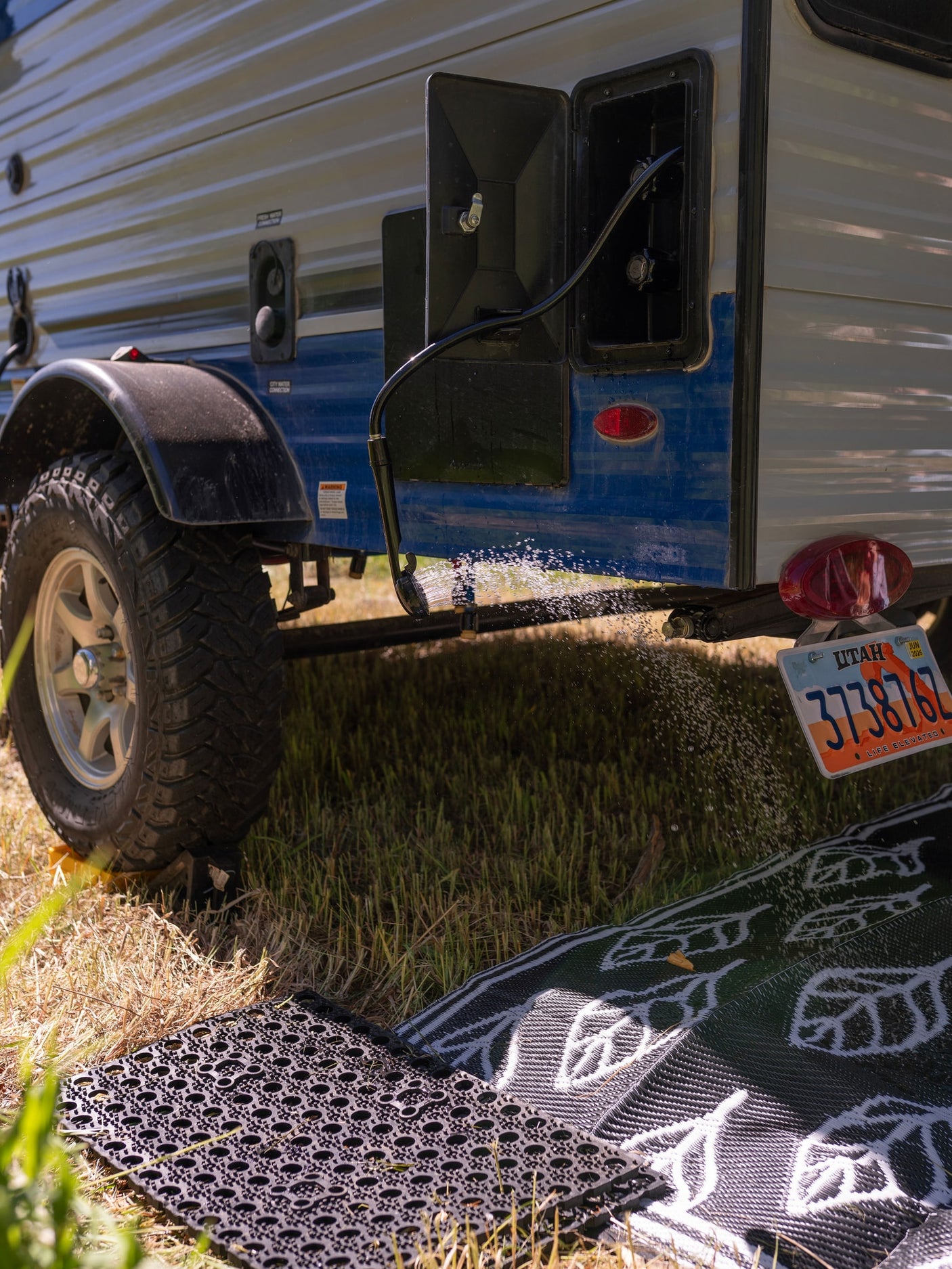 RV rental with an outdoor shower spraying water onto ground mats. A Utah-plated camper is seen in a grassy setting.