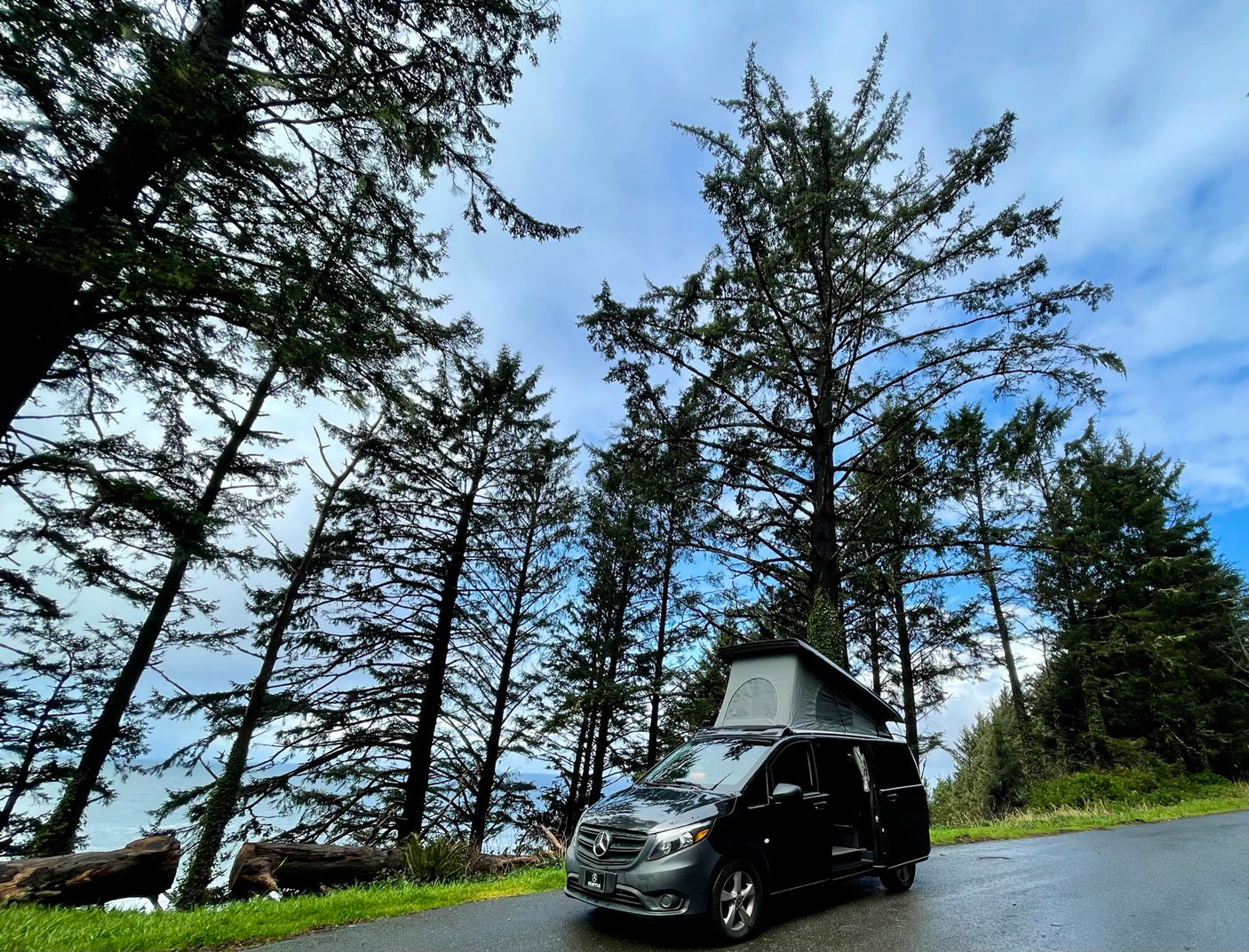 Black Mercedes campervan RV rental with pop-top extended, parked on a road lined with tall evergreen trees overlooking the ocean.