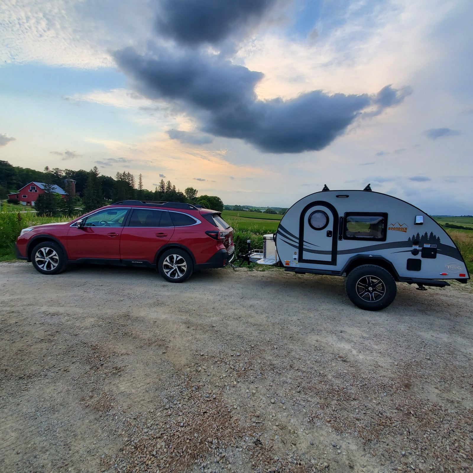 Red Subaru Outback towing a grey teardrop RV rental on a gravel road with a rural farm in the distance under a cloudy sky.