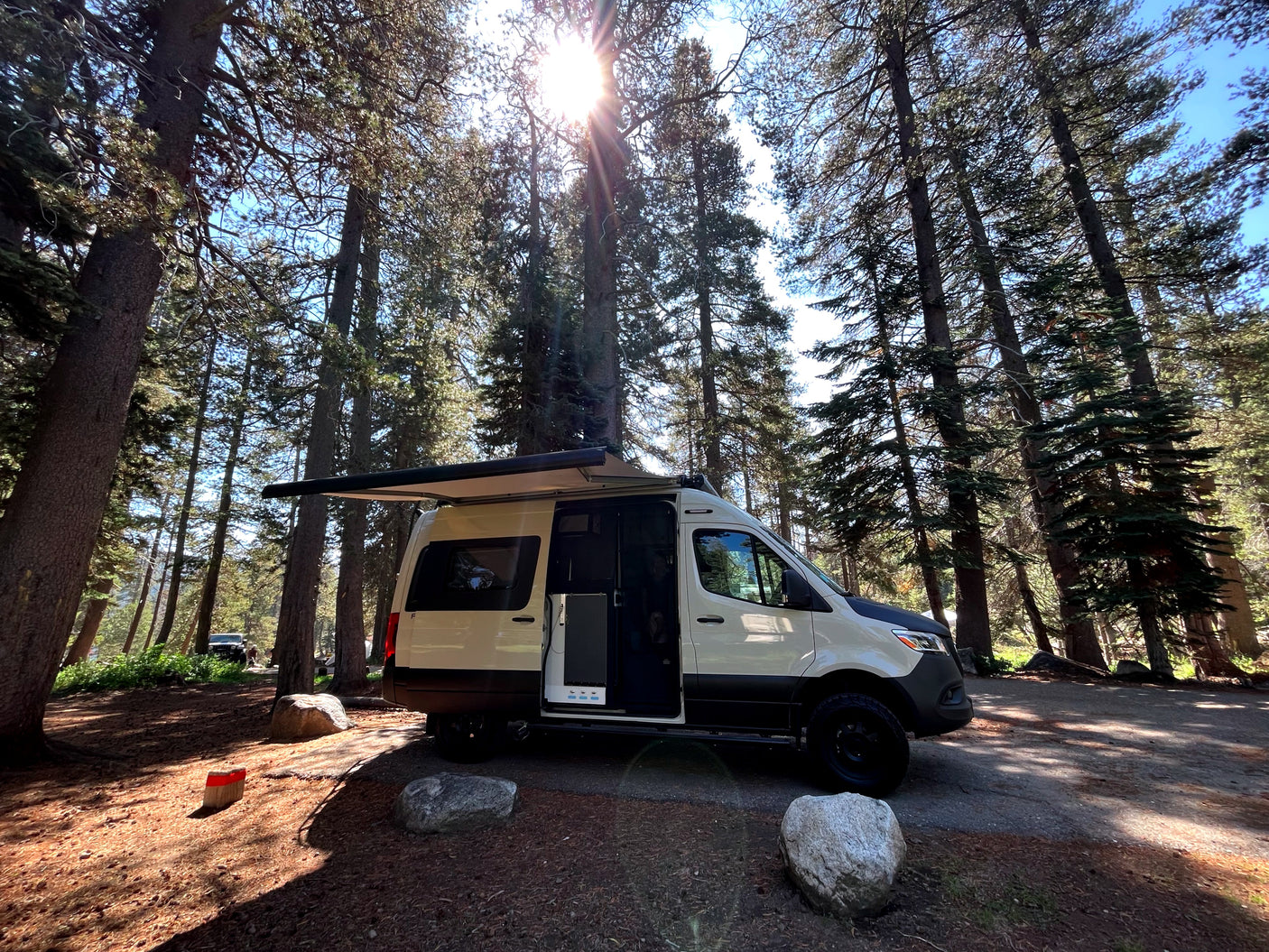 A white camper van rv rental with an extended awning is parked at a sunlit forest campsite, ready for outdoor living.