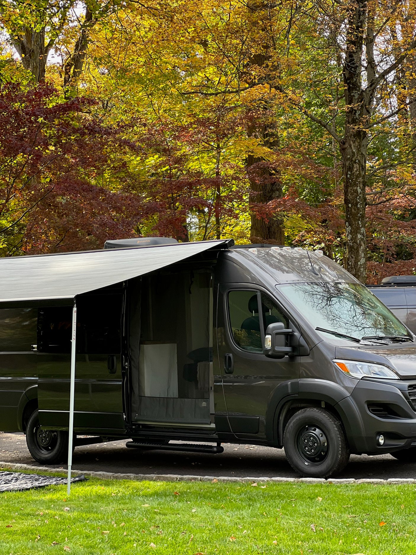 Dark grey RV rental camper van with open door and extended awning, parked among vibrant autumn trees.