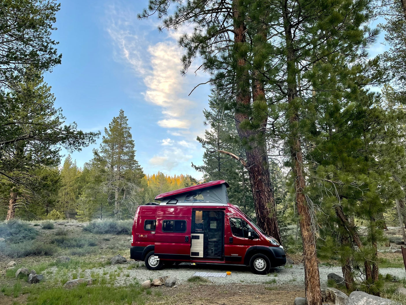 parked against an arid landscape with a bright rainbow. A dog watches from inside.|A red camper van (RV rental) is parked among tall pine trees at a scenic forest campsite with its pop-top extended