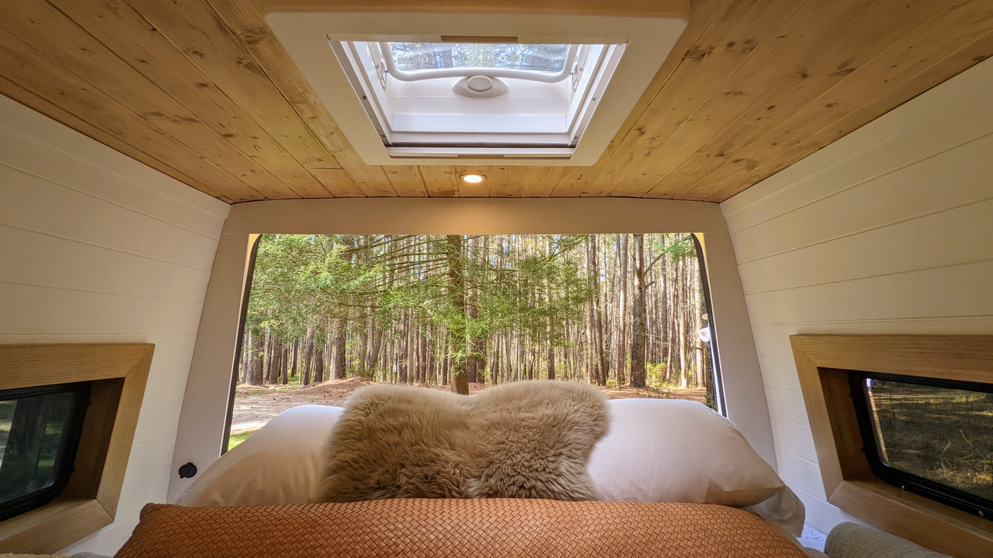 Inside an RV rental: wooden ceiling, skylight, and comfy bed facing a dense forest. Escape to nature's tranquility.